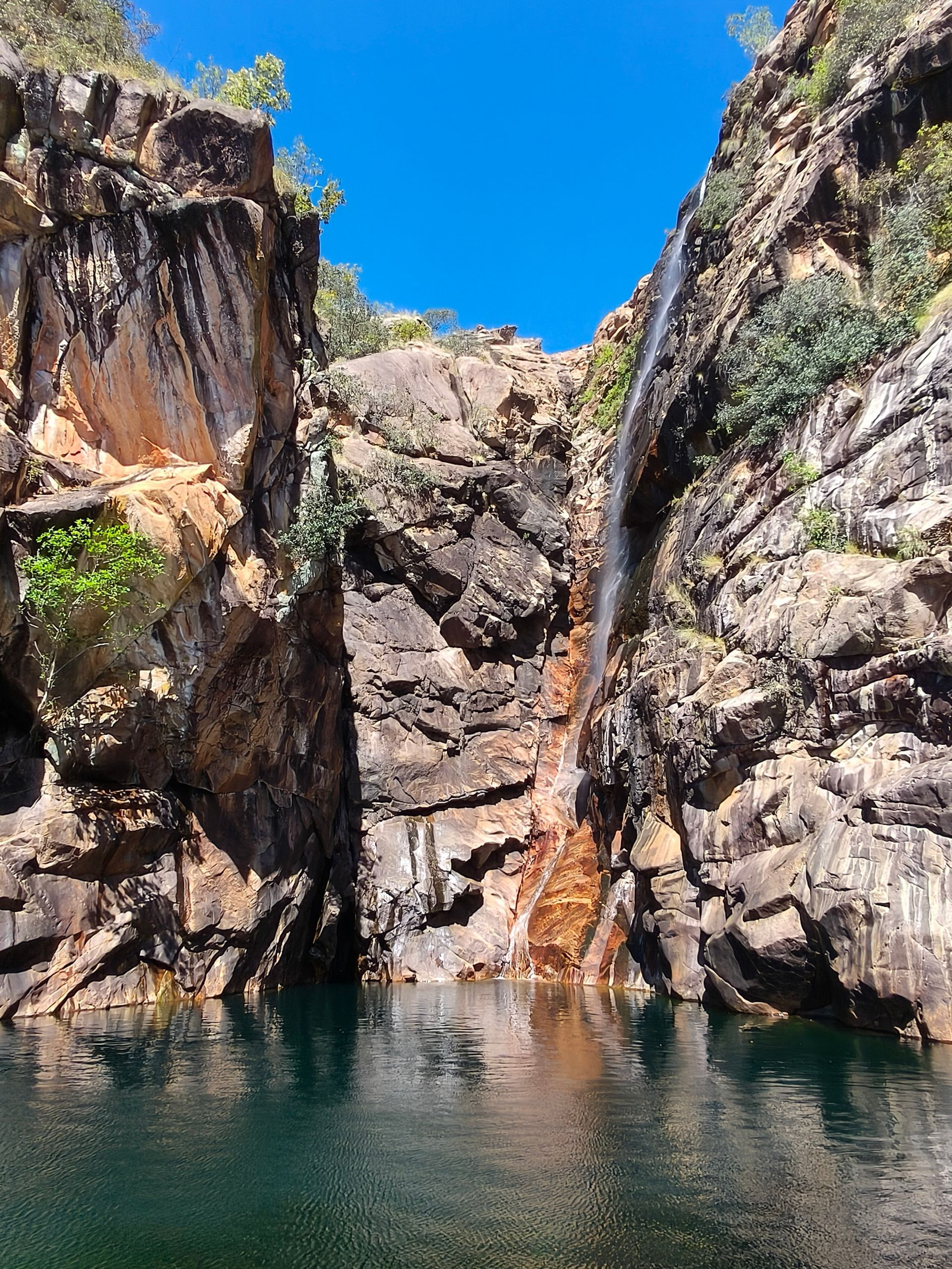 Waterfall cascading into a clear pool, flanked by rugged brown rock cliffs and a bright blue sky  — NT Driver Guide - Escorted Tours & Transfers in Darwin City, NT