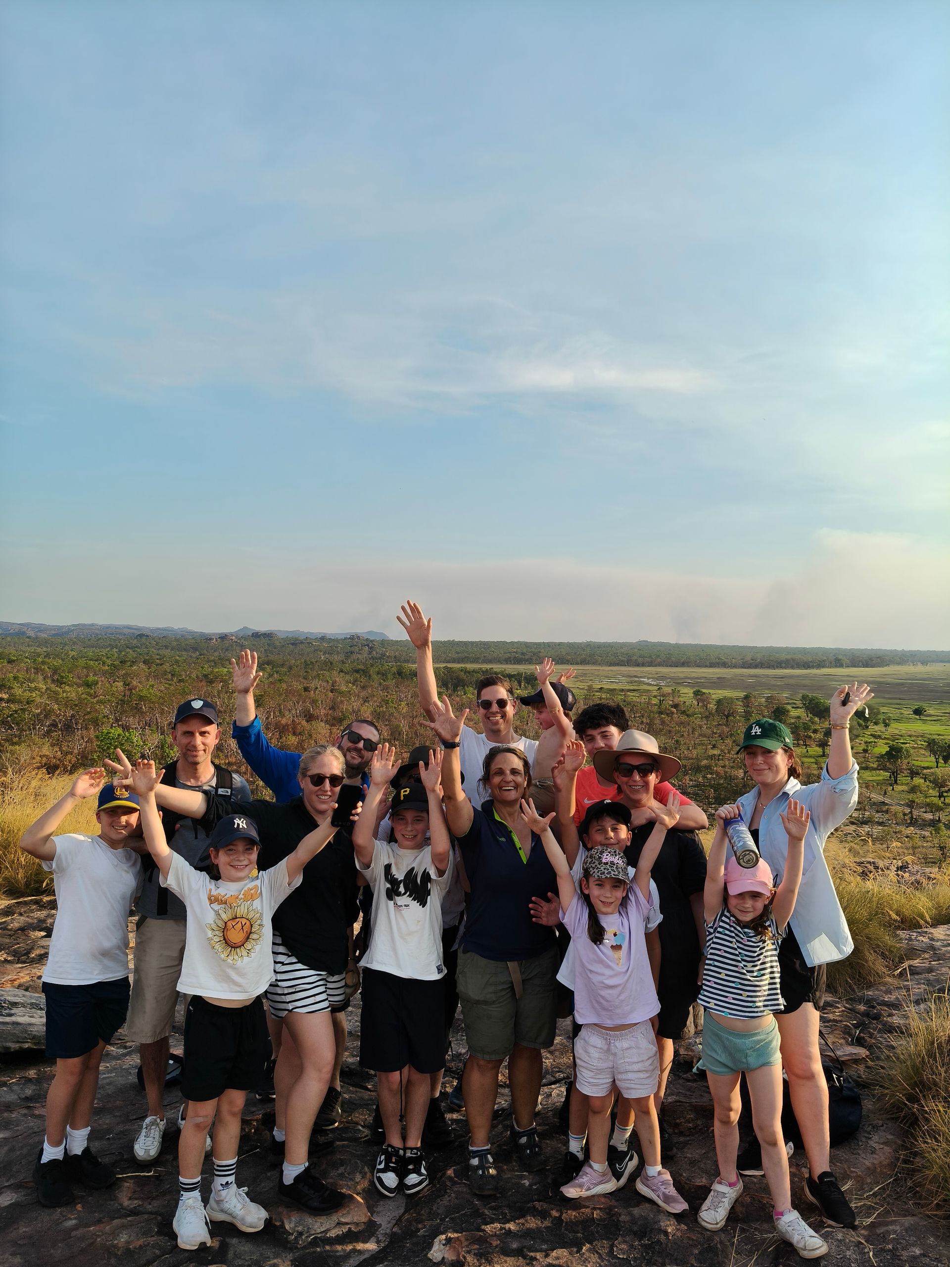 Group of people with arms raised on a rocky outcrop, with trees and a blue sky in the background — NT Driver Guide - Escorted Tours & Transfers in Darwin City, NT