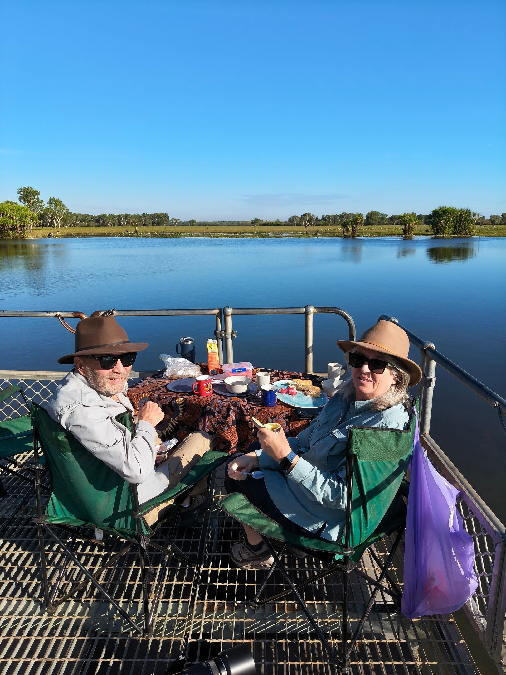 Two people in hats and sunglasses enjoy a meal outdoors on a deck overlooking calm water — NT Driver Guide - Escorted Tours & Transfers in Darwin City, NT