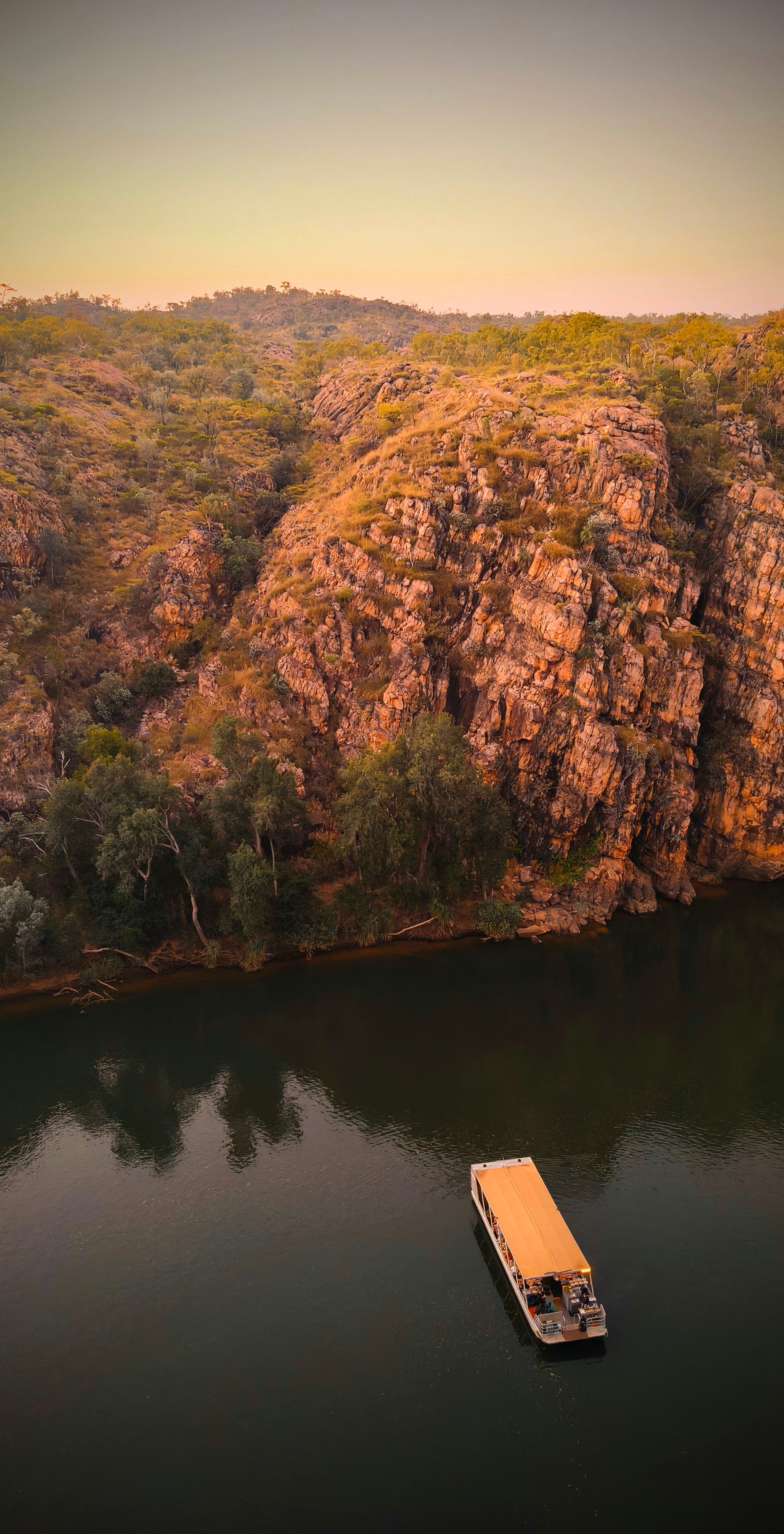 Boat on dark water near a rocky cliffside covered with brown and green vegetation — NT Driver Guide - Escorted Tours & Transfers in Darwin City, NT