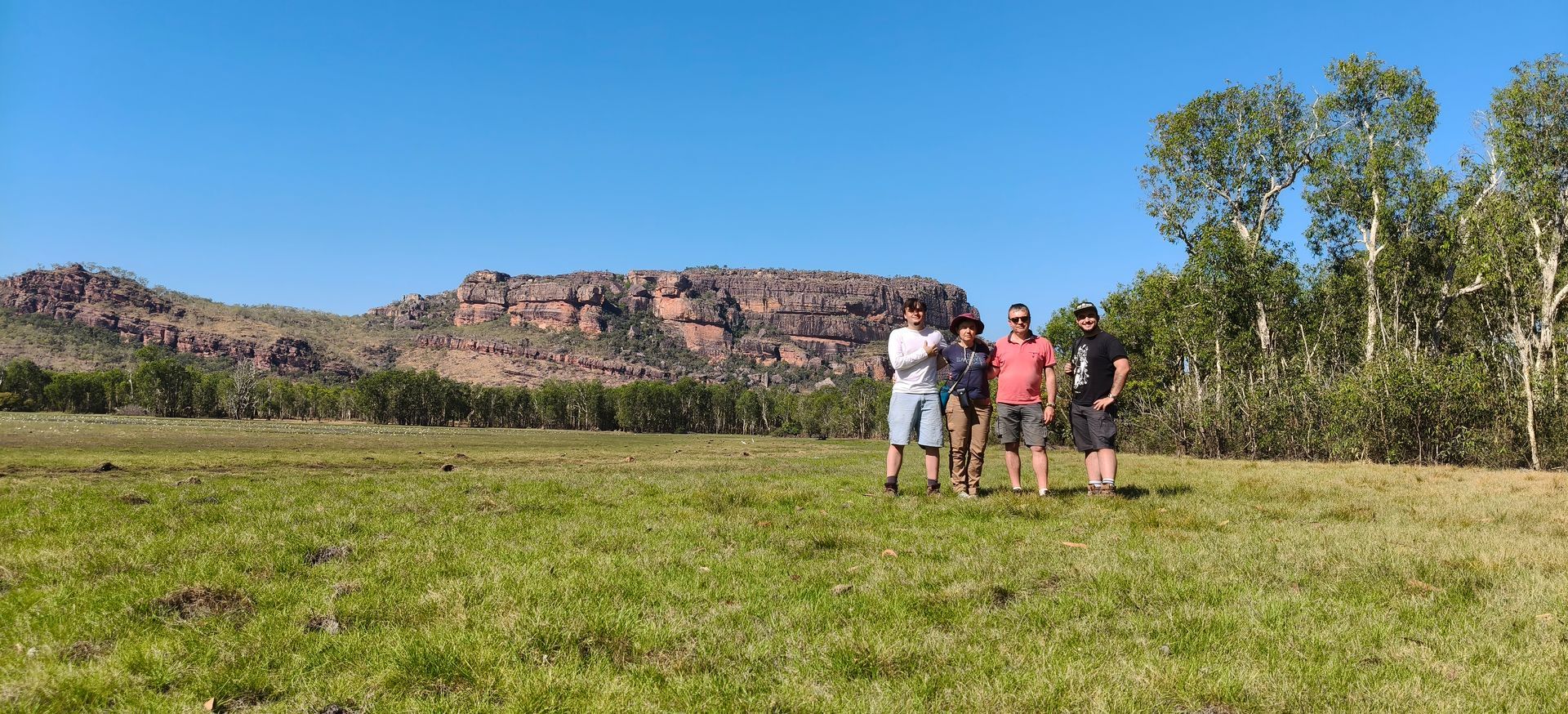 Four people standing in a grassy field with mountains in the background under a blue sky — NT Driver Guide - Escorted Tours & Transfers in Darwin City, NT