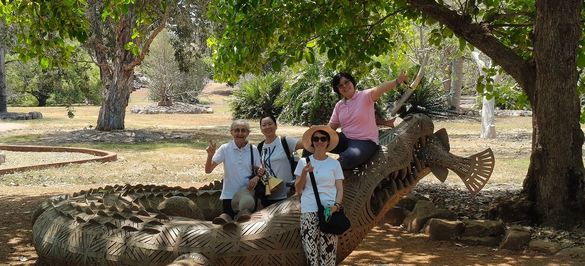 Four people pose on a metal sculpture of a dragon in a park, under trees. One person is sitting on it, arm raised — NT Driver Guide - Escorted Tours & Transfers in Darwin City, NT