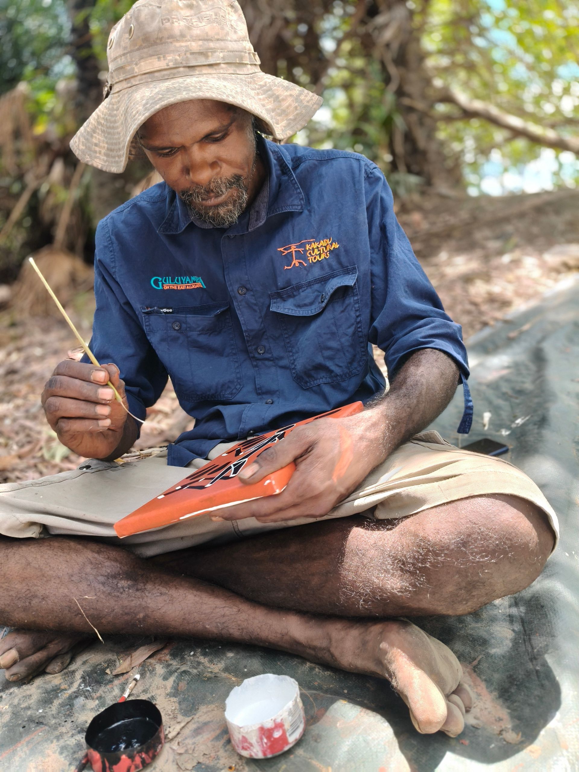 Man sitting outdoors, painting on an orange surface with a brush. He wears a hat and blue shirt — NT Driver Guide - Escorted Tours & Transfers in Darwin City, NT