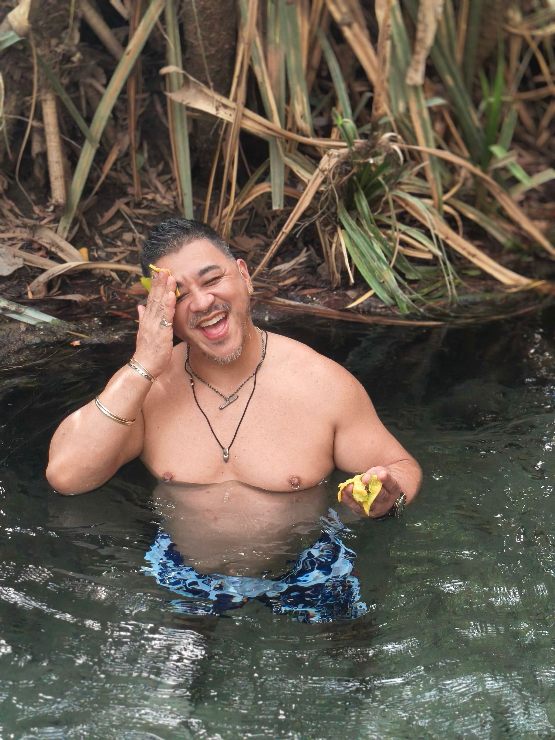 Man in water, smiling with hand on face. Wearing swim trunks, necklace. Natural setting with plants — NT Driver Guide - Escorted Tours & Transfers in Darwin City, NT