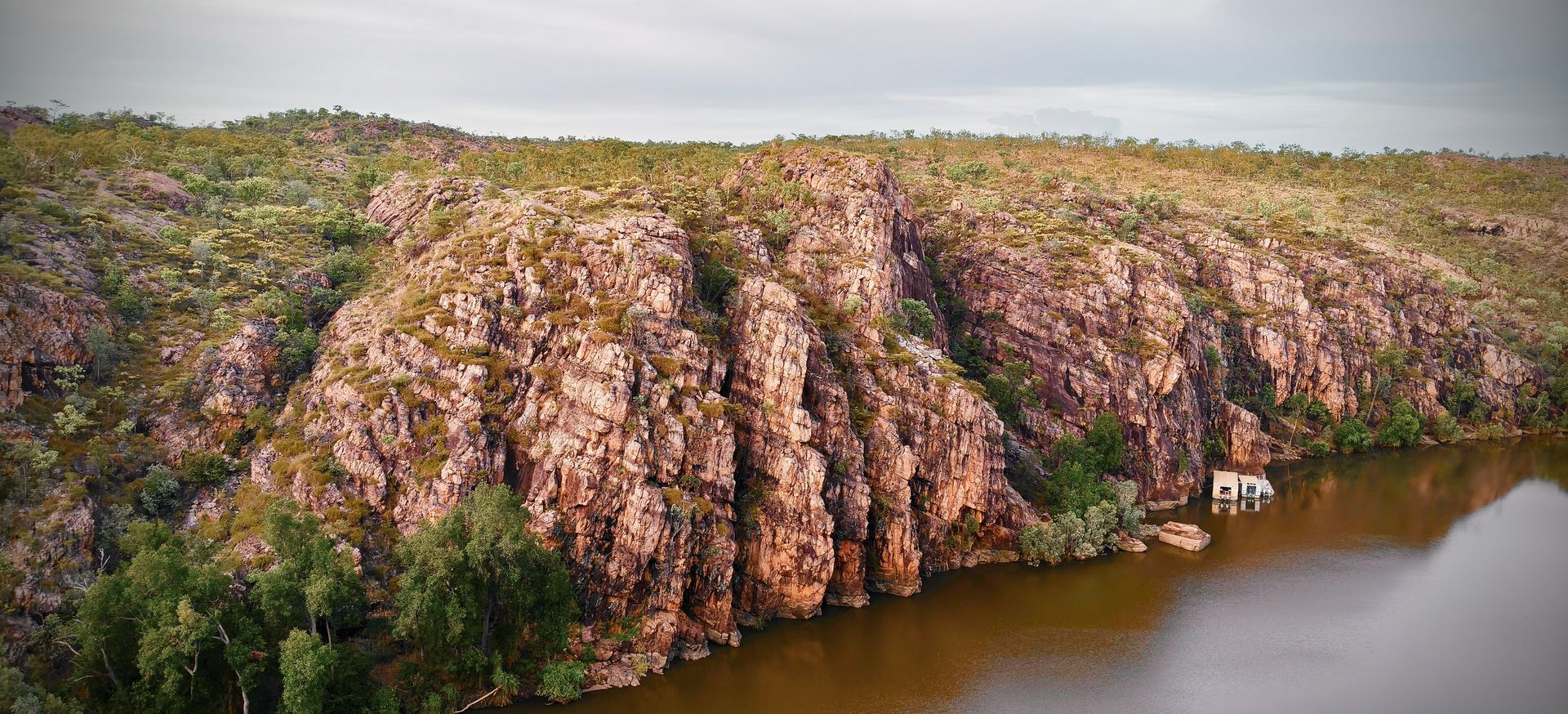 Rocky cliffside with vegetation overlooking a murky lake, small white building at water's edge — NT Driver Guide - Escorted Tours & Transfers in Darwin City, NT