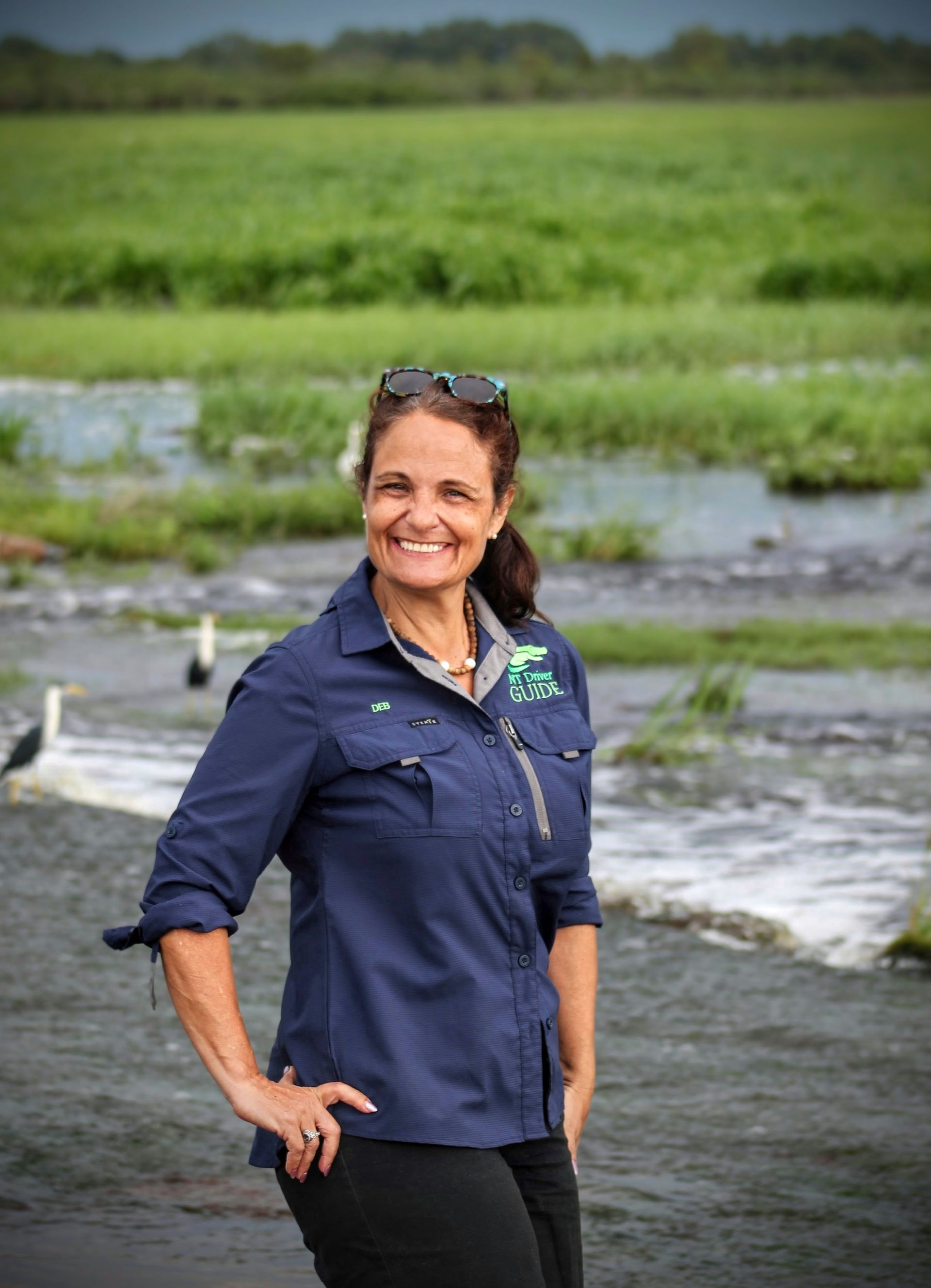Woman in blue shirt gestures on a balcony, smiling, with a lush green backdrop — NT Driver Guide - Escorted Tours & Transfers in Darwin City, NT