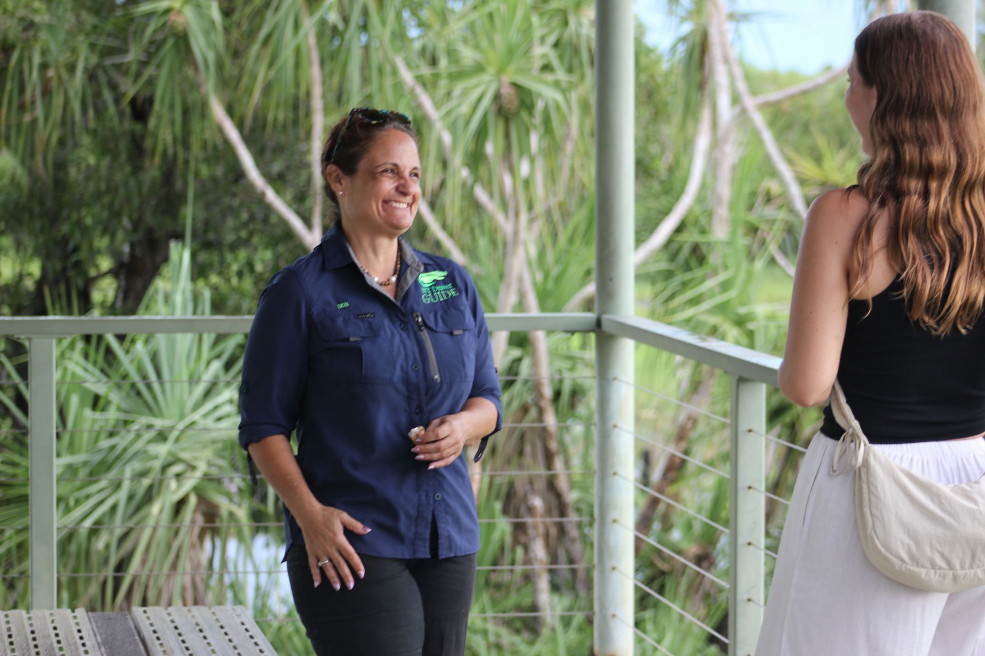 Woman in blue shirt smiles, talking on a balcony with another person, surrounded by green foliage — NT Driver Guide - Escorted Tours & Transfers in Darwin City, NT