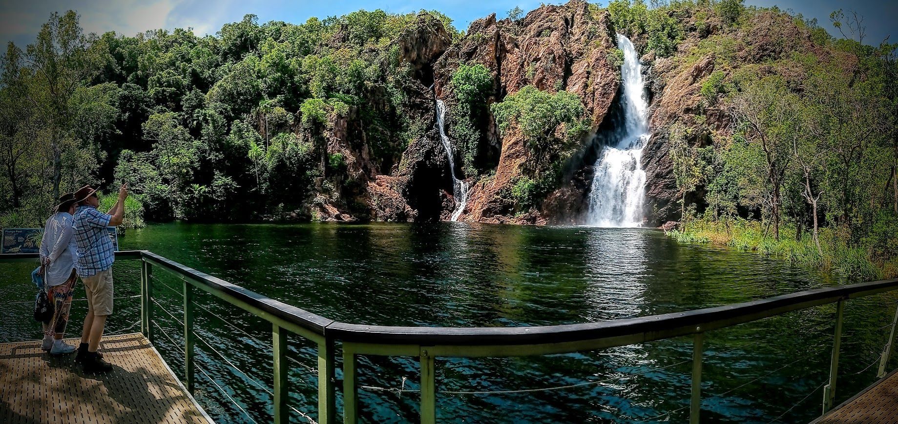 A person stands on a pier overlooking a waterfall and a lake surrounded by trees — NT Driver Guide - Escorted Tours & Transfers in Darwin City, NT