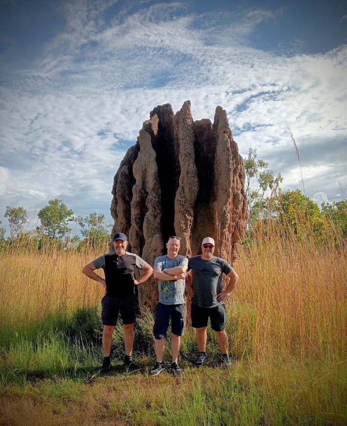 Three men stand near a large termite mound in an open, grassy field under a cloudy sky — NT Driver Guide - Escorted Tours & Transfers in Darwin City, NT