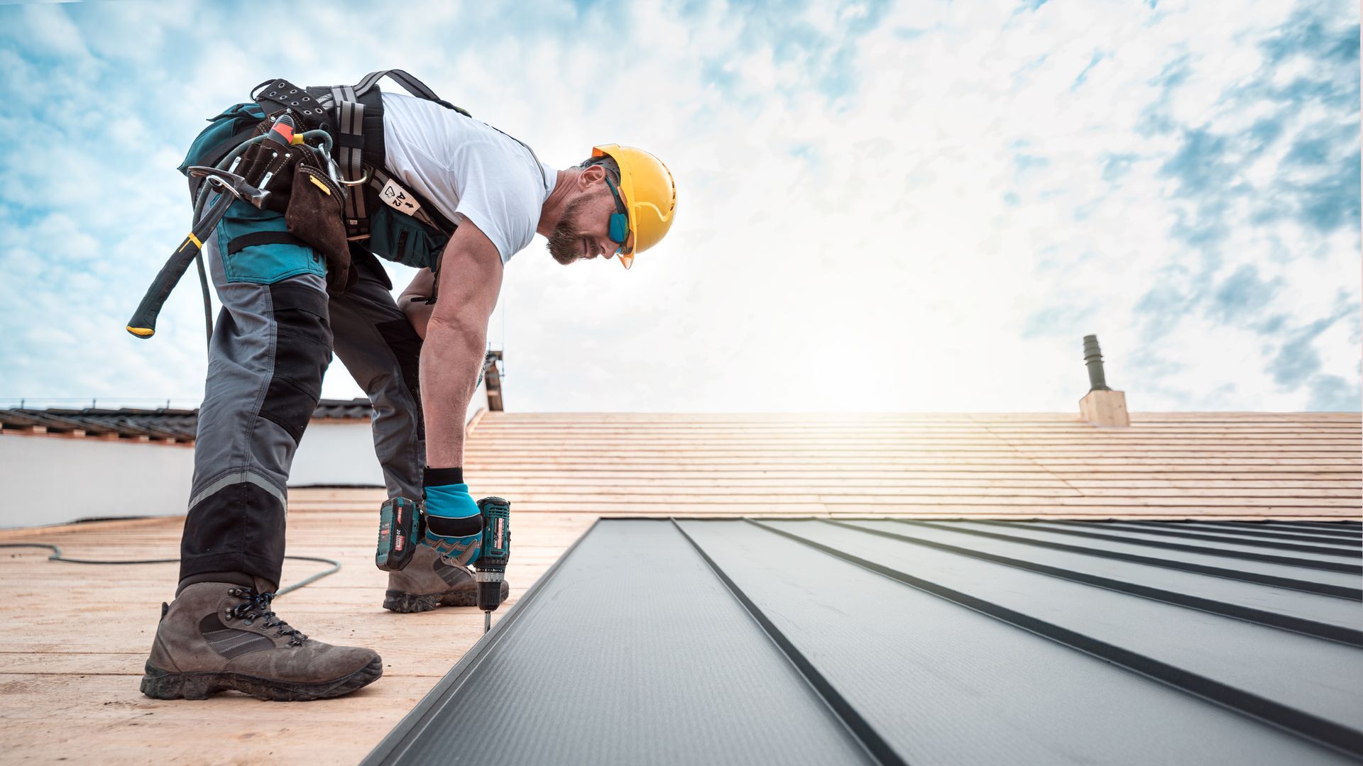 A male roofer with a safety harness is anchoring metal roofing to a wooden base with a screwdriver.