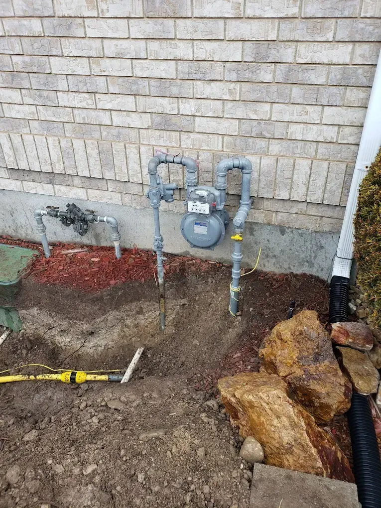 Gas meter and pipes against a brick wall, in a partially dug area, with landscaping materials.
