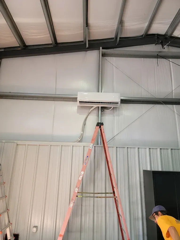 Air conditioner mounted on a metal wall, with a ladder beneath it. A person is working.