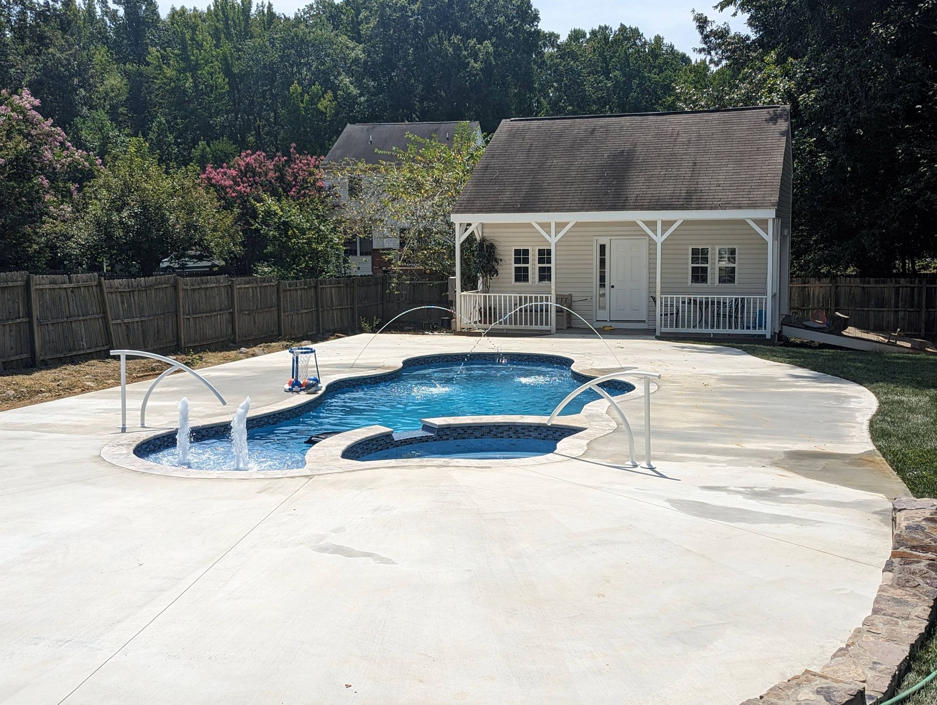 A backyard swimming pool with fountains and an attached spa, set in a patio area in front of a small cottage-style building.