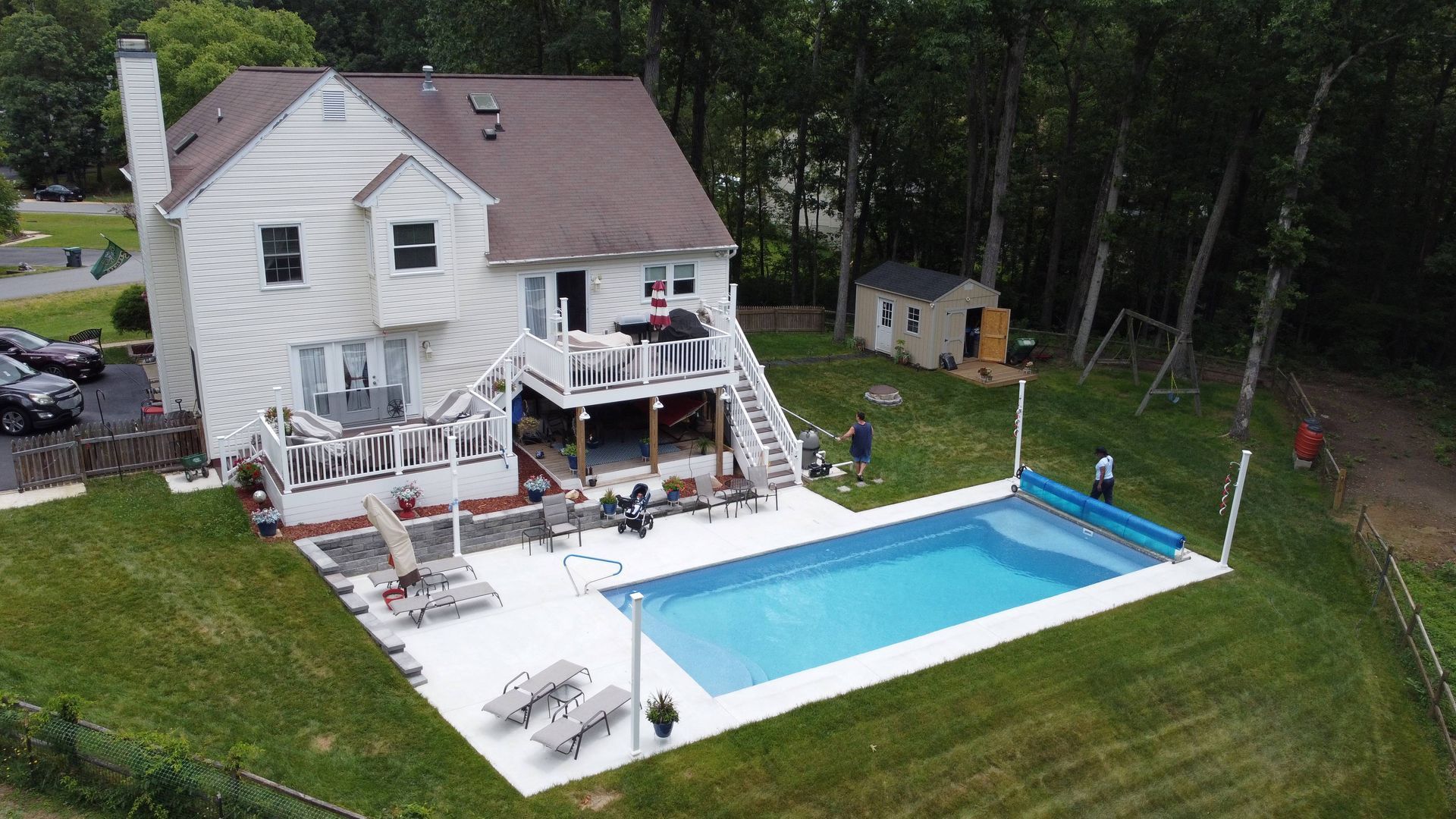 Aerial view of a white house with a multi-level deck, patio, and rectangular swimming pool in a grassy backyard.