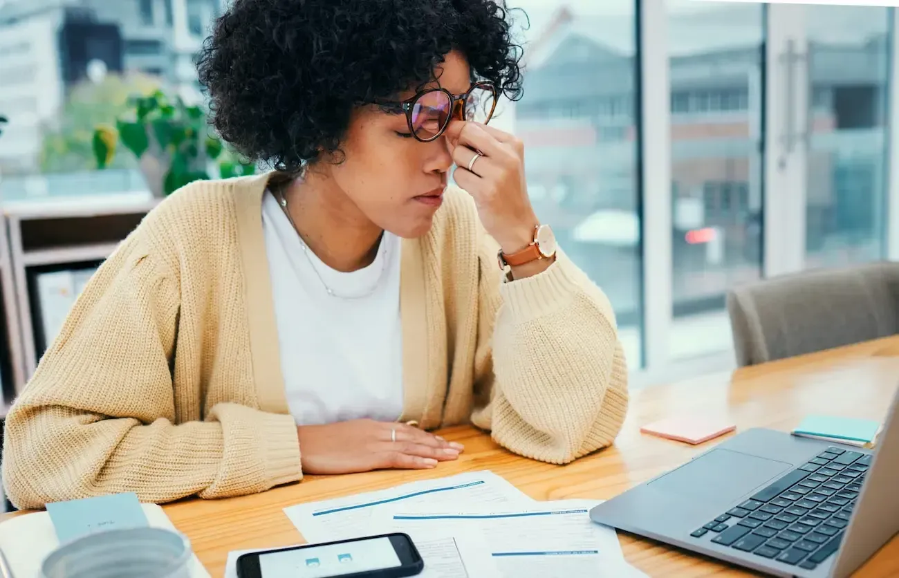 A woman is sitting at a desk with a laptop and rubbing her eyes.