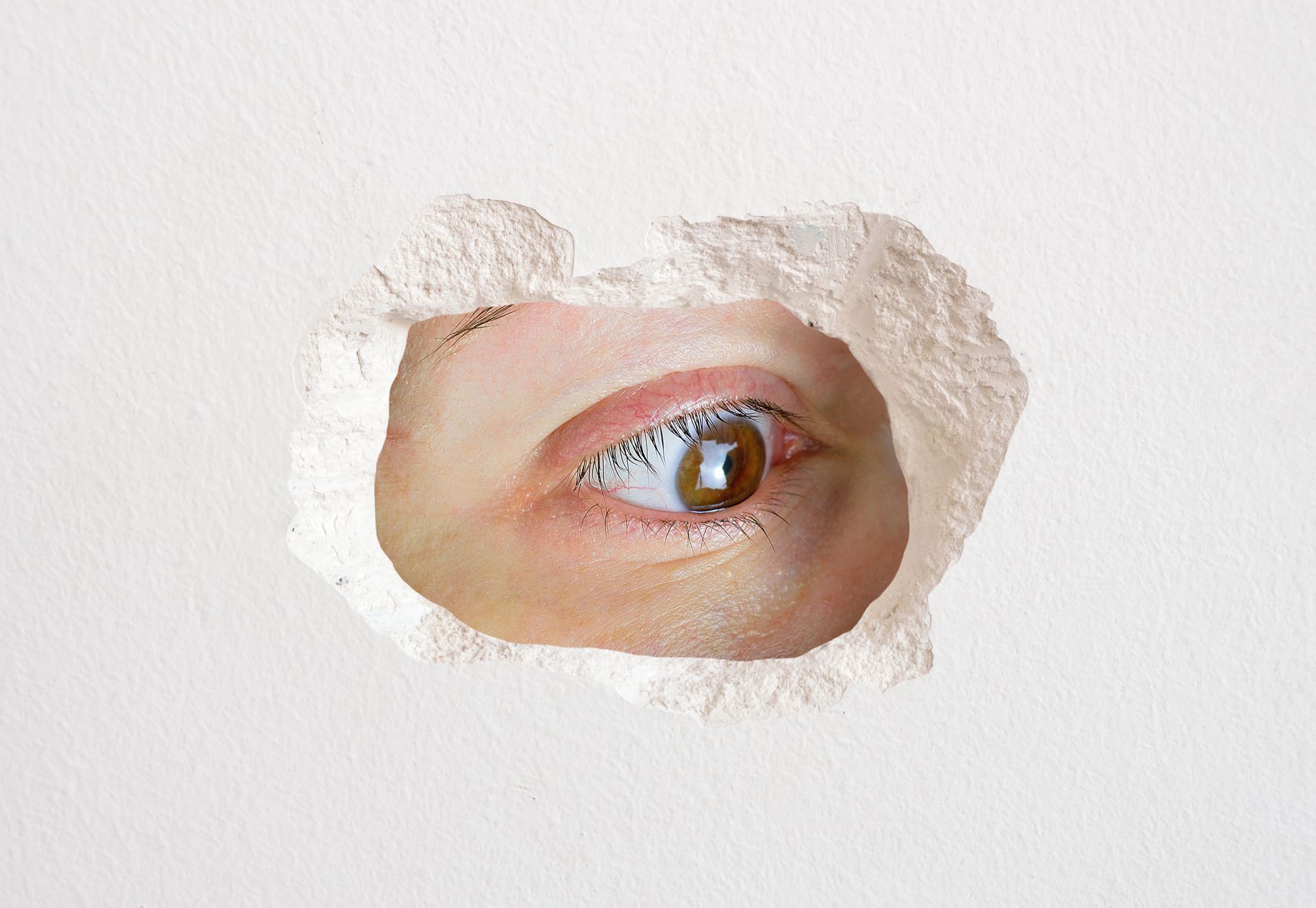 Eye peeking through a hole in a white wall.