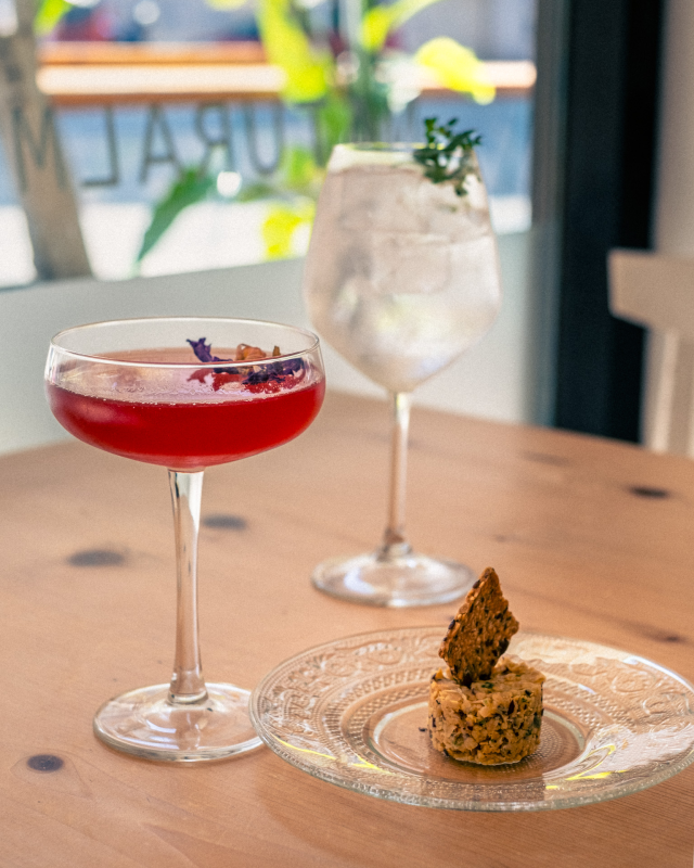 Due cocktail e un piccolo pasticcino su un tavolo di legno vicino a una finestra. Un drink è rosso, l'altro è trasparente.