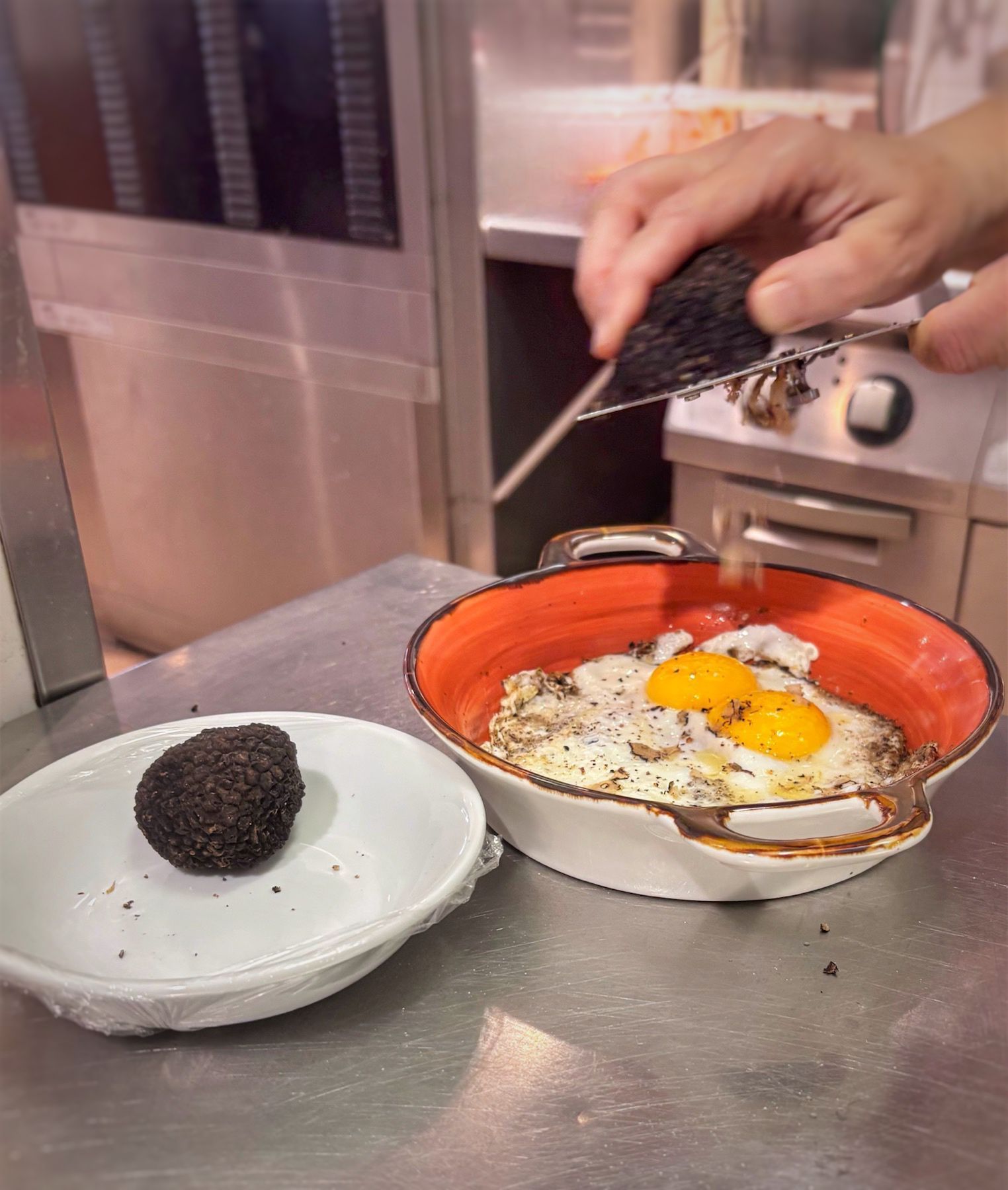 Persona che grattugia il tartufo nero sulle uova fritte in un piatto colorato. Un tartufo è appoggiato su un piattino.