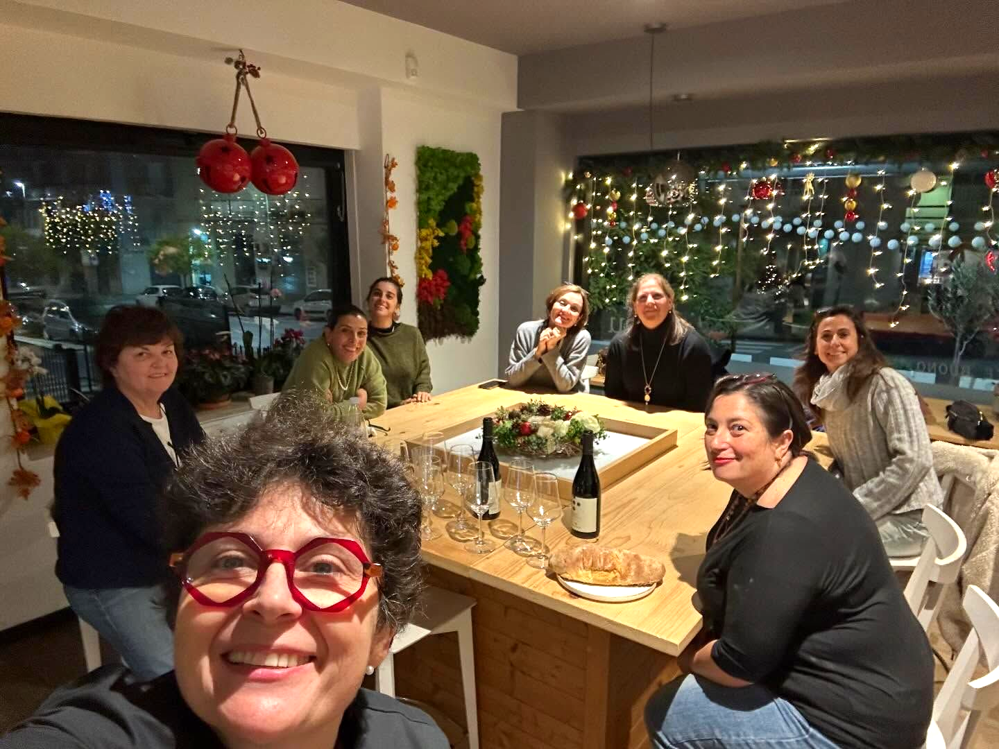 Un gruppo di donne è riunito attorno a un tavolo di legno, sorridenti, all'interno, vicino a una grande finestra. Sono visibili decorazioni natalizie.