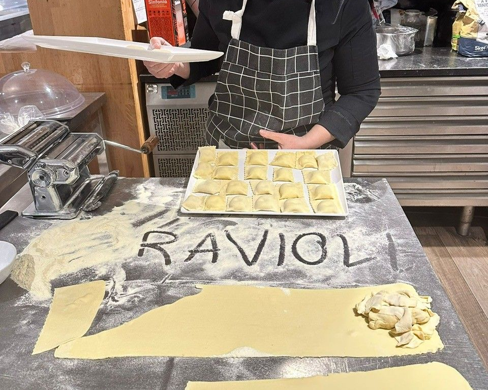 Persona che prepara i ravioli in cucina. Tiene in mano un piatto, con un vassoio di ravioli ripieni e pasta sul tavolo infarinato.
