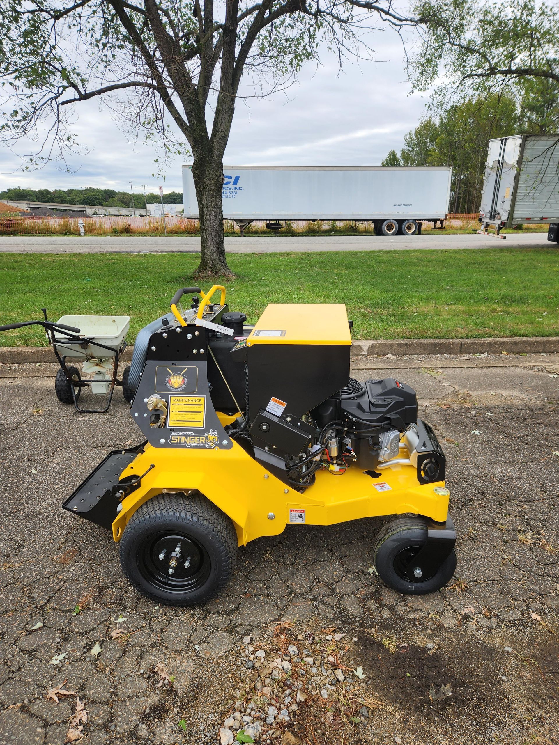 A yellow lawn mower is parked in a parking lot next to a tree.