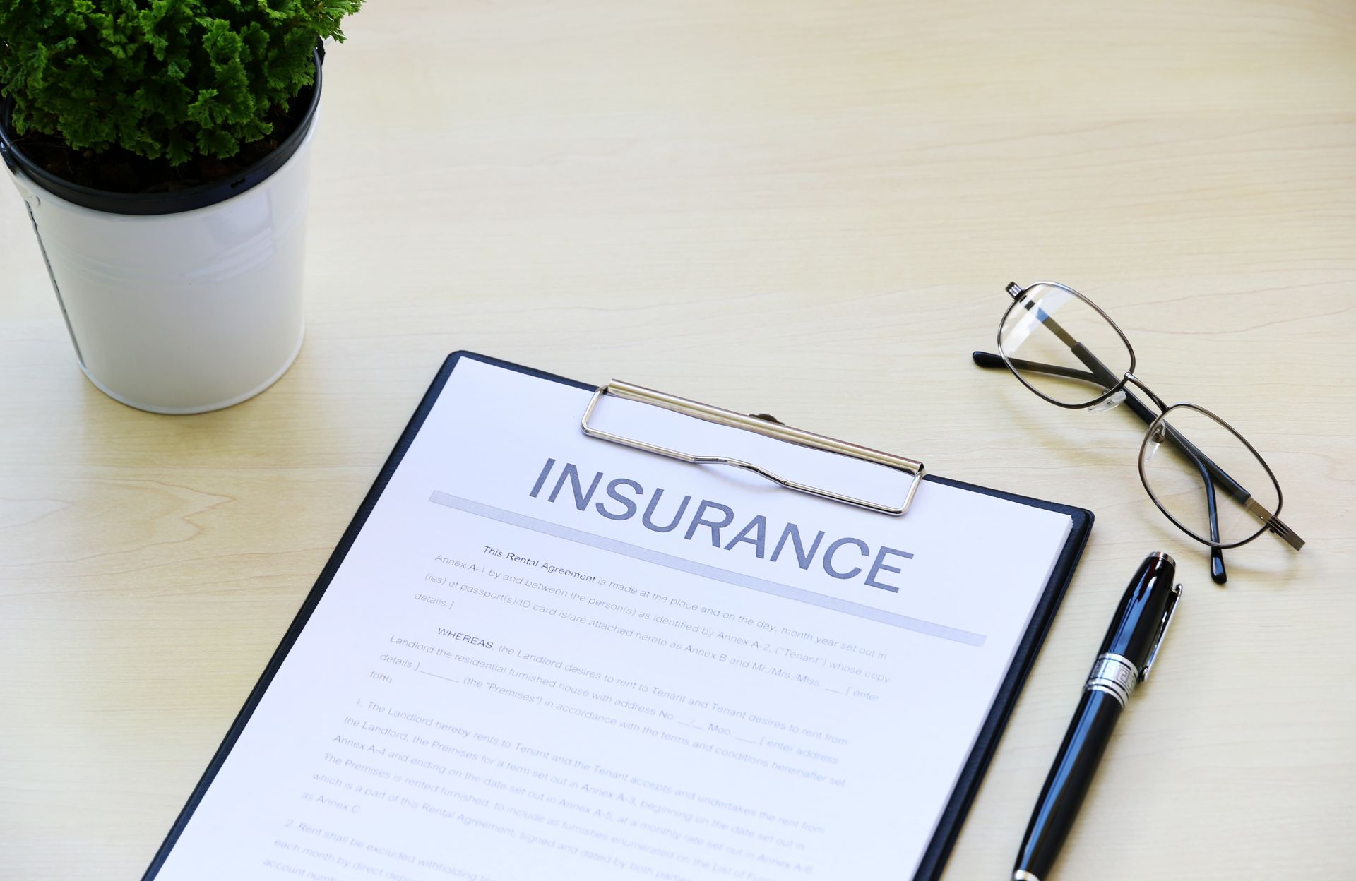 Clipboard with “INSURANCE” form, pen, glasses, and potted plant on a desk.