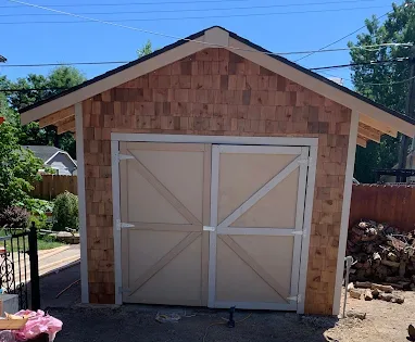 Wooden garage with light beige doors and cedar shingle siding, under a bright blue sky.