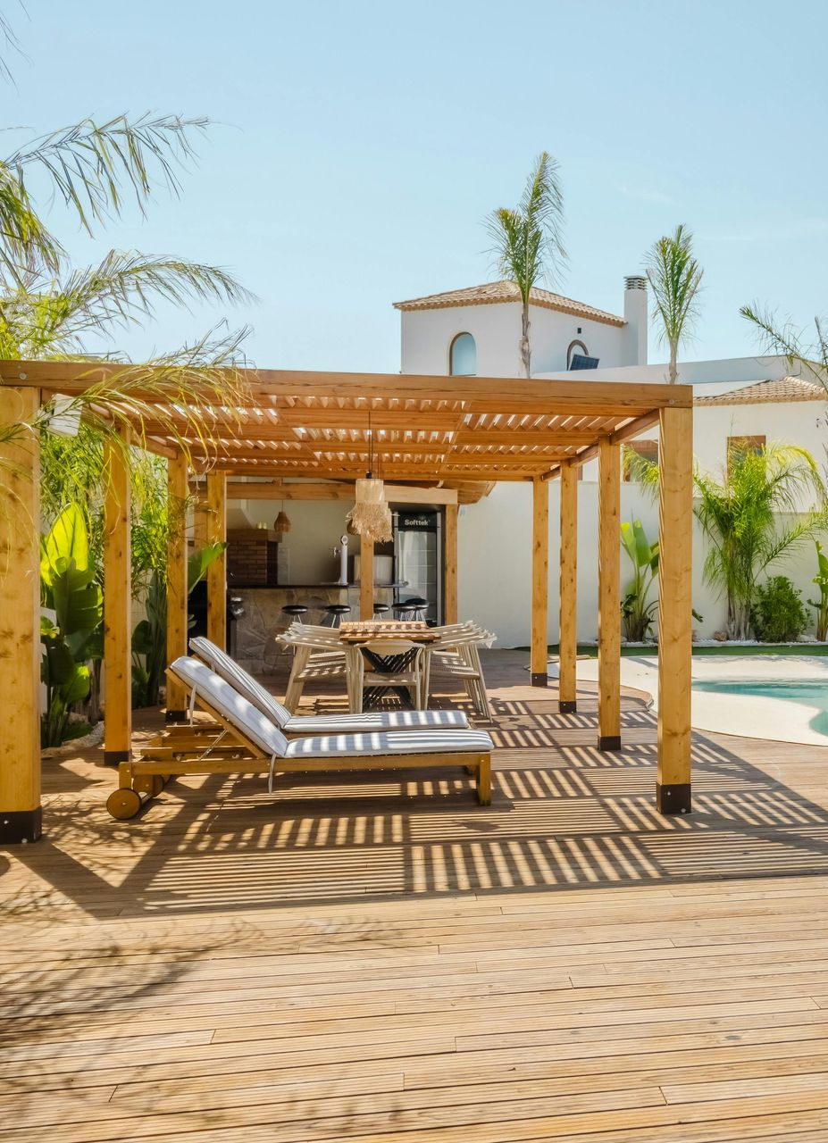 Wooden pergola over lounge chairs, dining area, near pool. Sunny day with shadows.