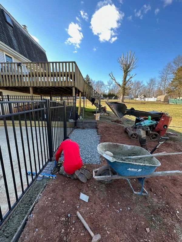 A worker laying pavers next to a black fence and a deck; equipment nearby, sunny day.
