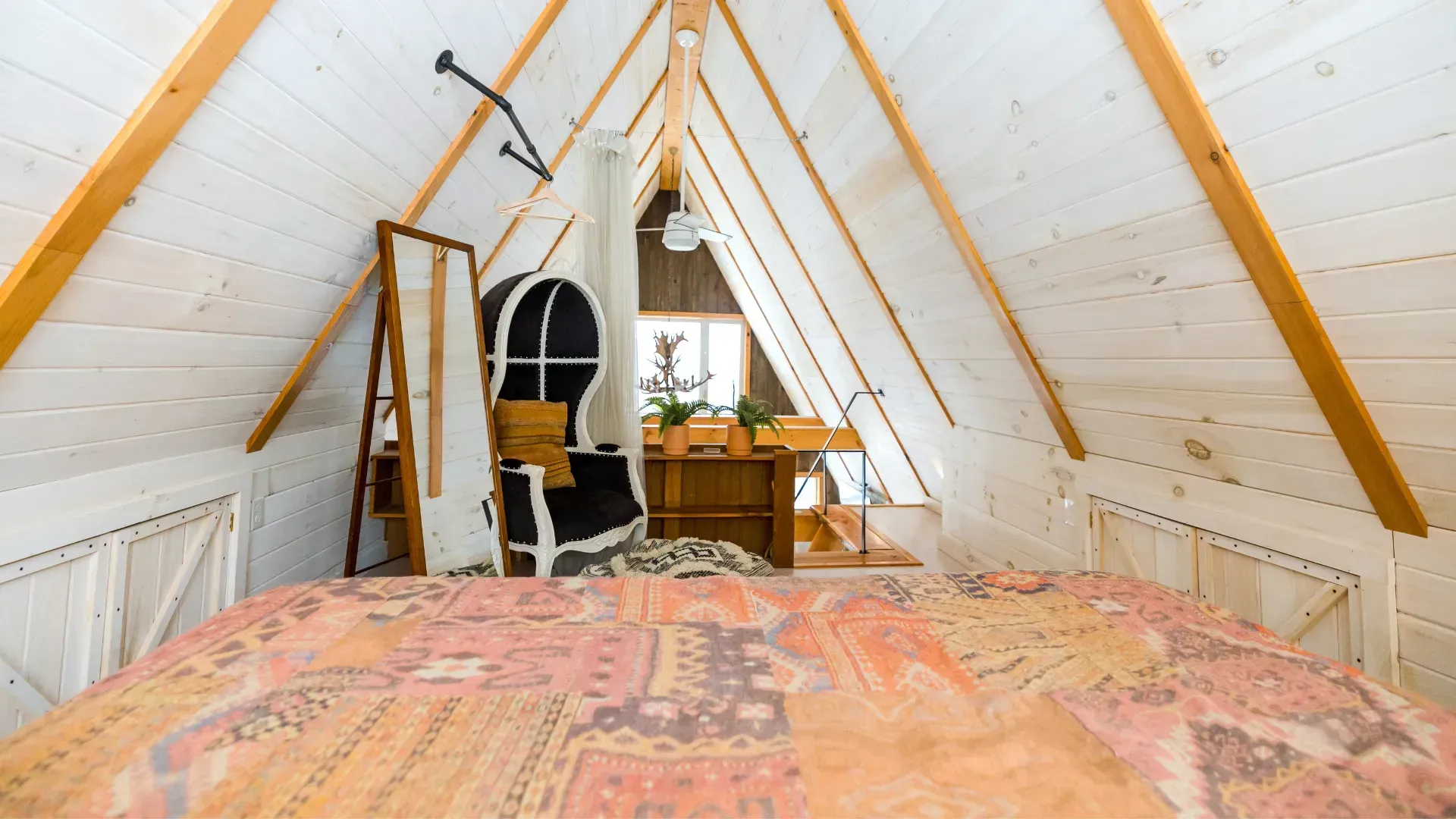 Loft bedroom with a bed, chair, and wood furniture against a white-paneled wall and ceiling.