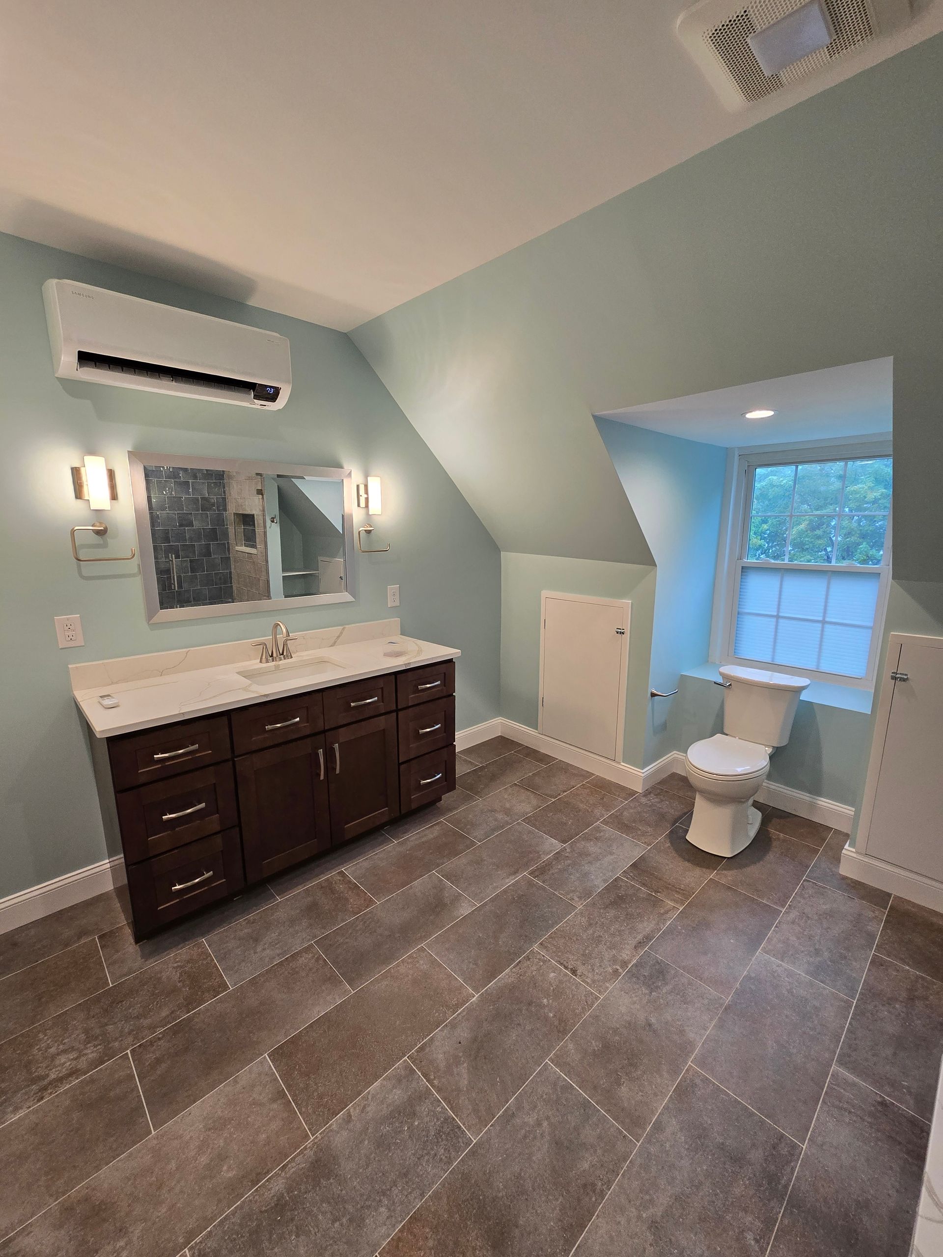 A bathroom with a toilet , sink , mirror and air conditioner.