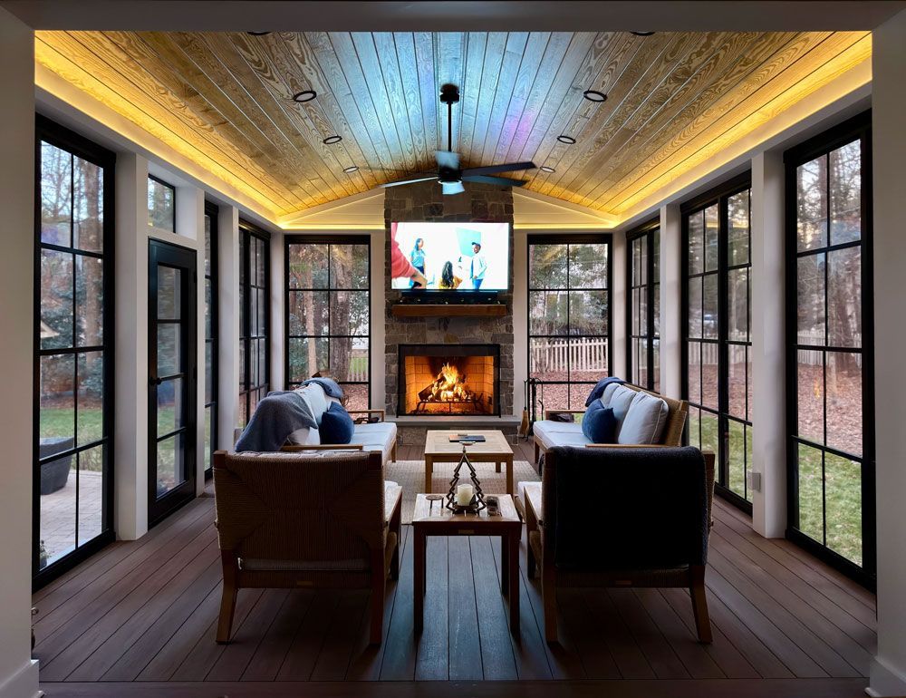 Cozy sunroom with fireplace, TV, and seating, surrounded by windows. Wooden floors, ceiling with color-changing lights.