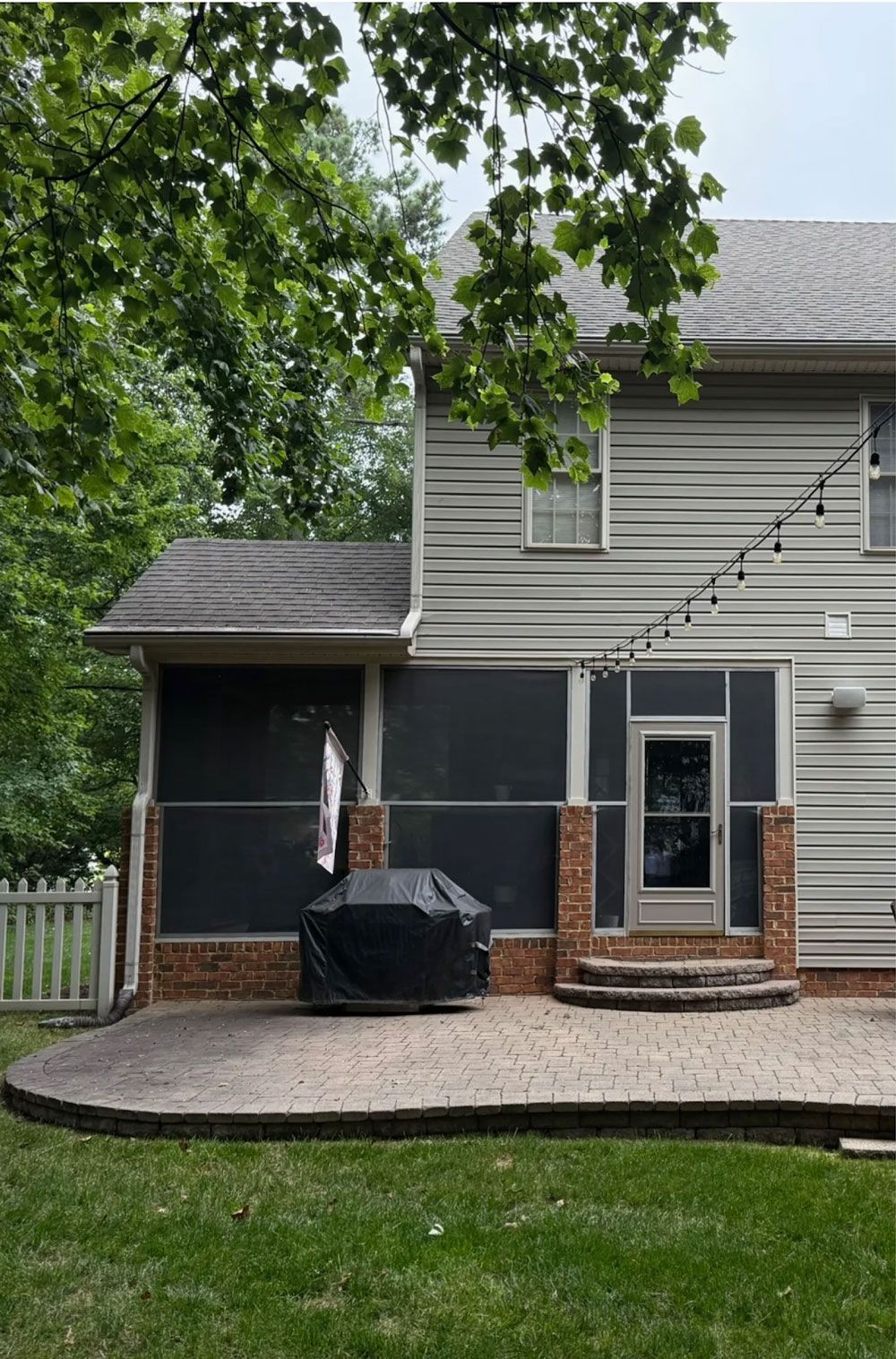 Backyard patio with brick columns, grill, screen-in porch, and string lights, surrounded by grass and trees.