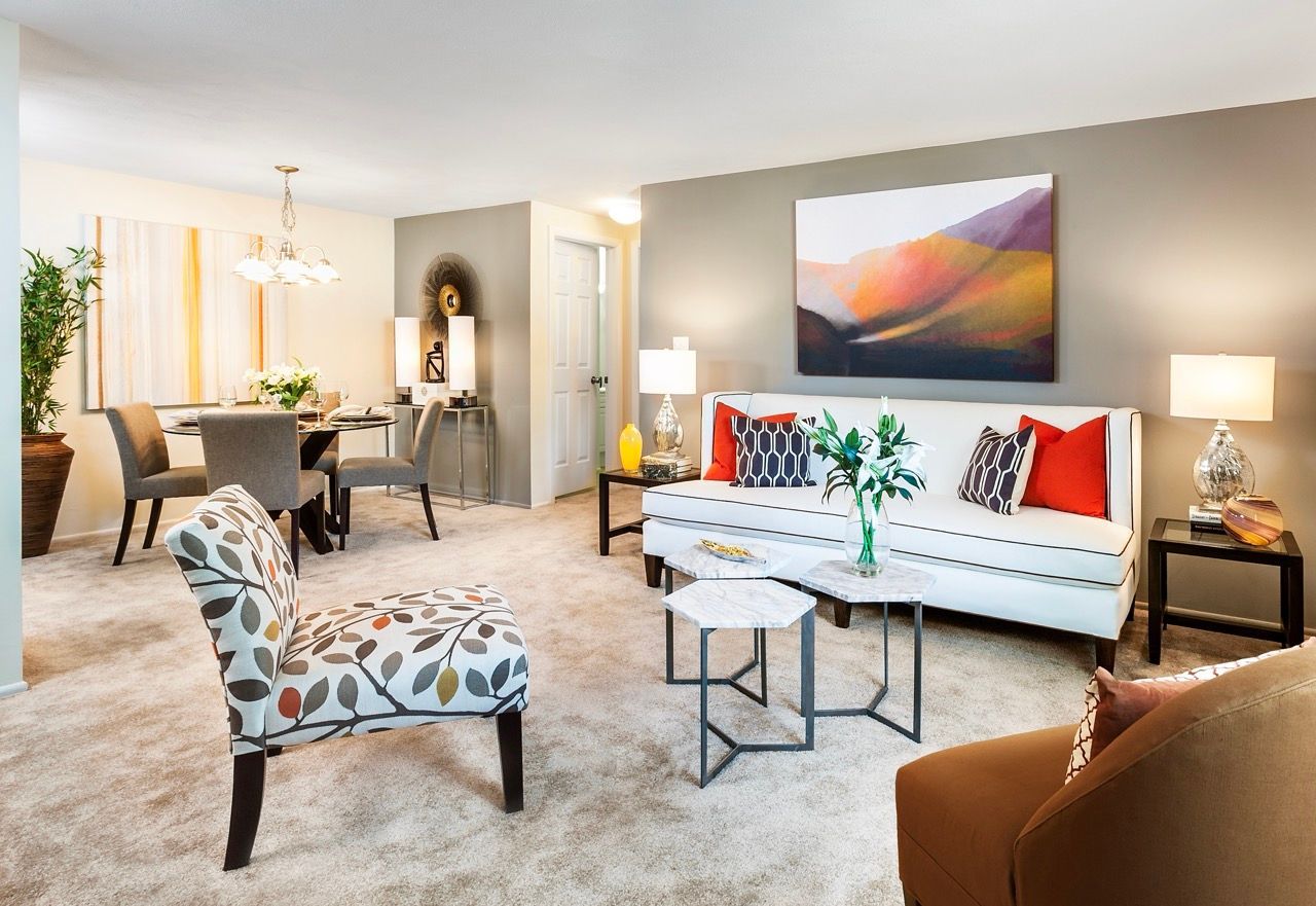 Bright living room in a furnished apartment with a white sofa, patterned chair, and glass coffee tables.