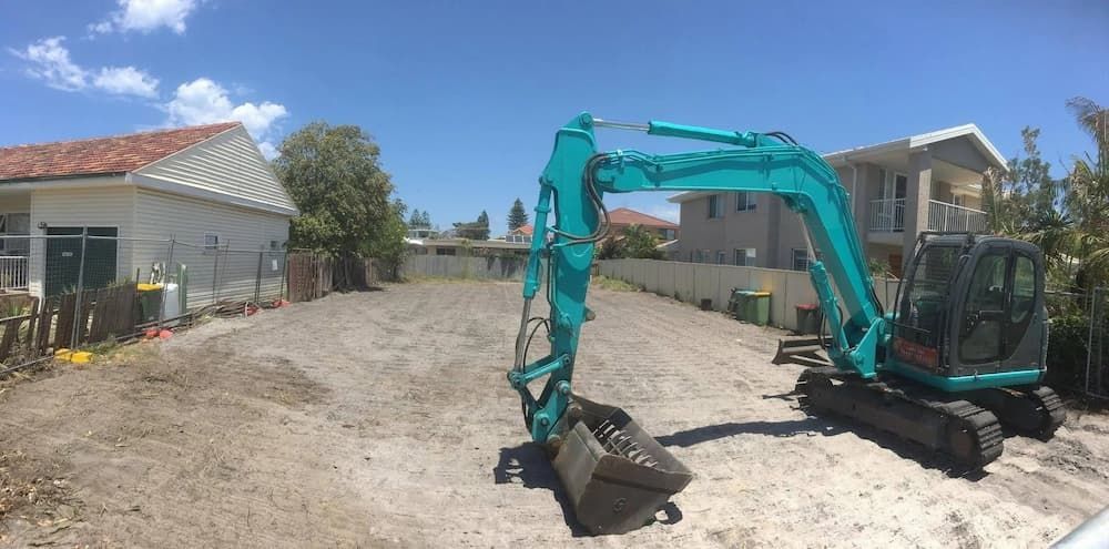 A Turquoise Excavator Sits on a Cleared, Sandy Lot Between Two Houses Under a Bright Blue Sky