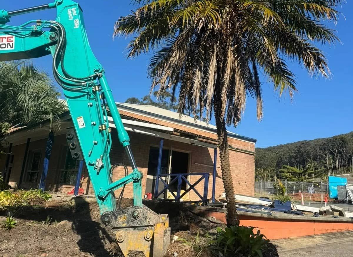 A Blue Excavator Is Parked in Front of A Building Next to A Palm Tree — Central Coast Complete Demolition & Tree Service in Toukley, NSW