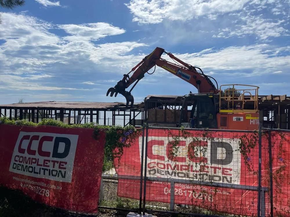 A Large Excavator Is Demolishing a Building Behind a Red Fence — Central Coast Complete Demolition & Tree Service in Buff Point, NSW