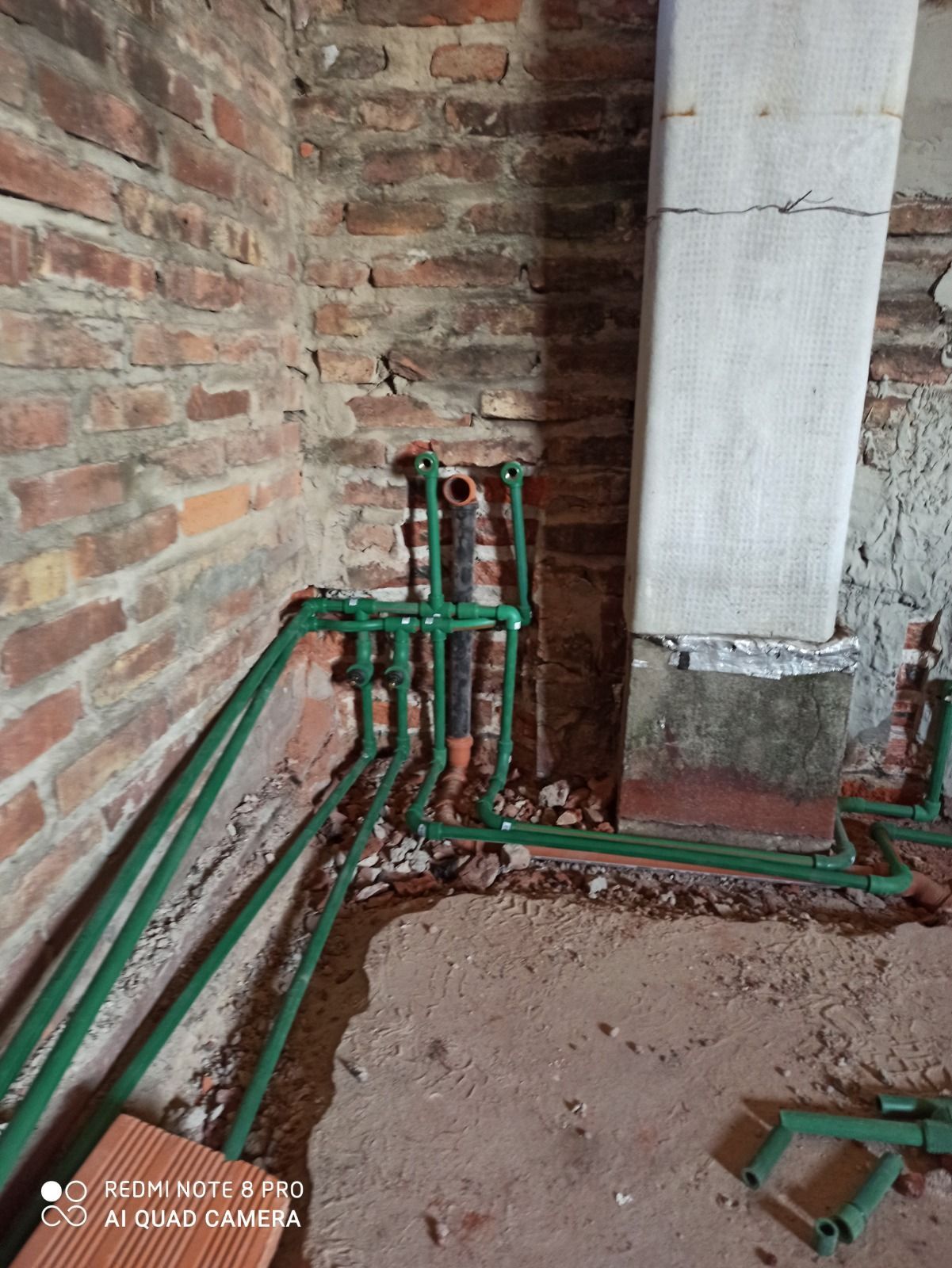 Tuberías verdes y rojas que recorren una pared de ladrillo en un rincón interior sin terminar.