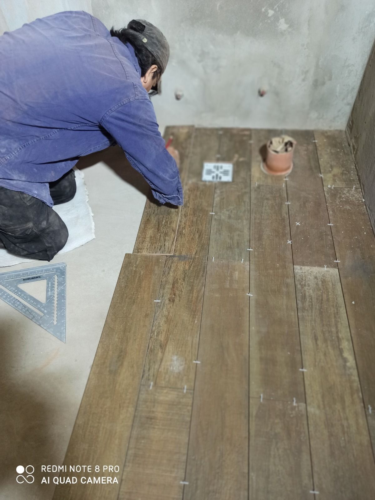 Una persona coloca tablones de madera en el suelo de una habitación con una pared de hormigón y herramientas cerca.