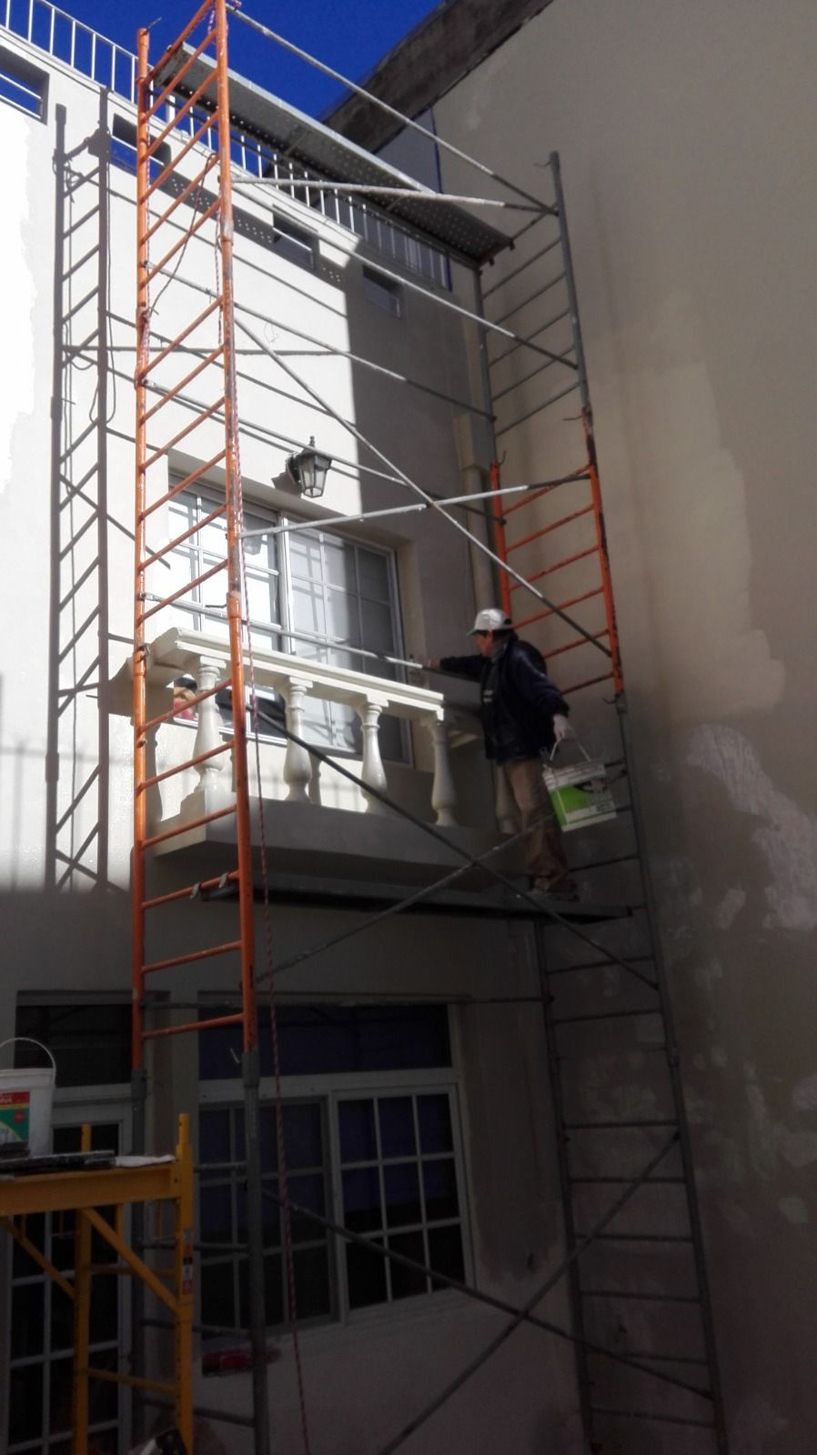 Un trabajador en un andamio alto junto a la pared blanca de un edificio, bañado por la luz del sol.
