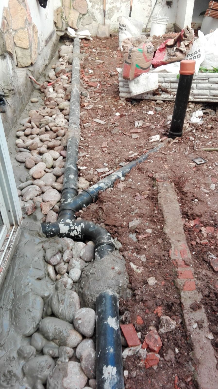 Zanja de tuberías dañada con tuberías de PVC negras y escombros junto a un muro en una zona de construcción.