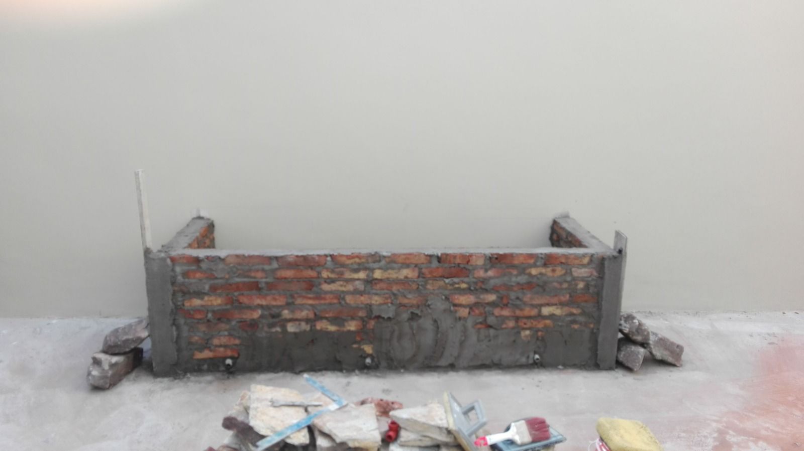 Pequeño muro de ladrillo en construcción contra una pared lisa, con escombros esparcidos por el suelo.