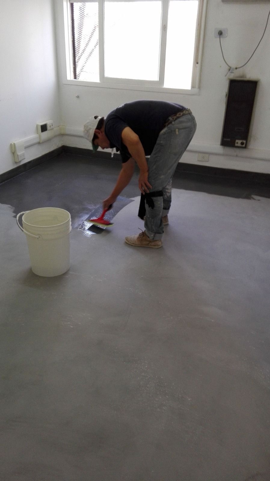 Una persona está fregando un suelo gris en una habitación con un cubo blanco y ventanas.