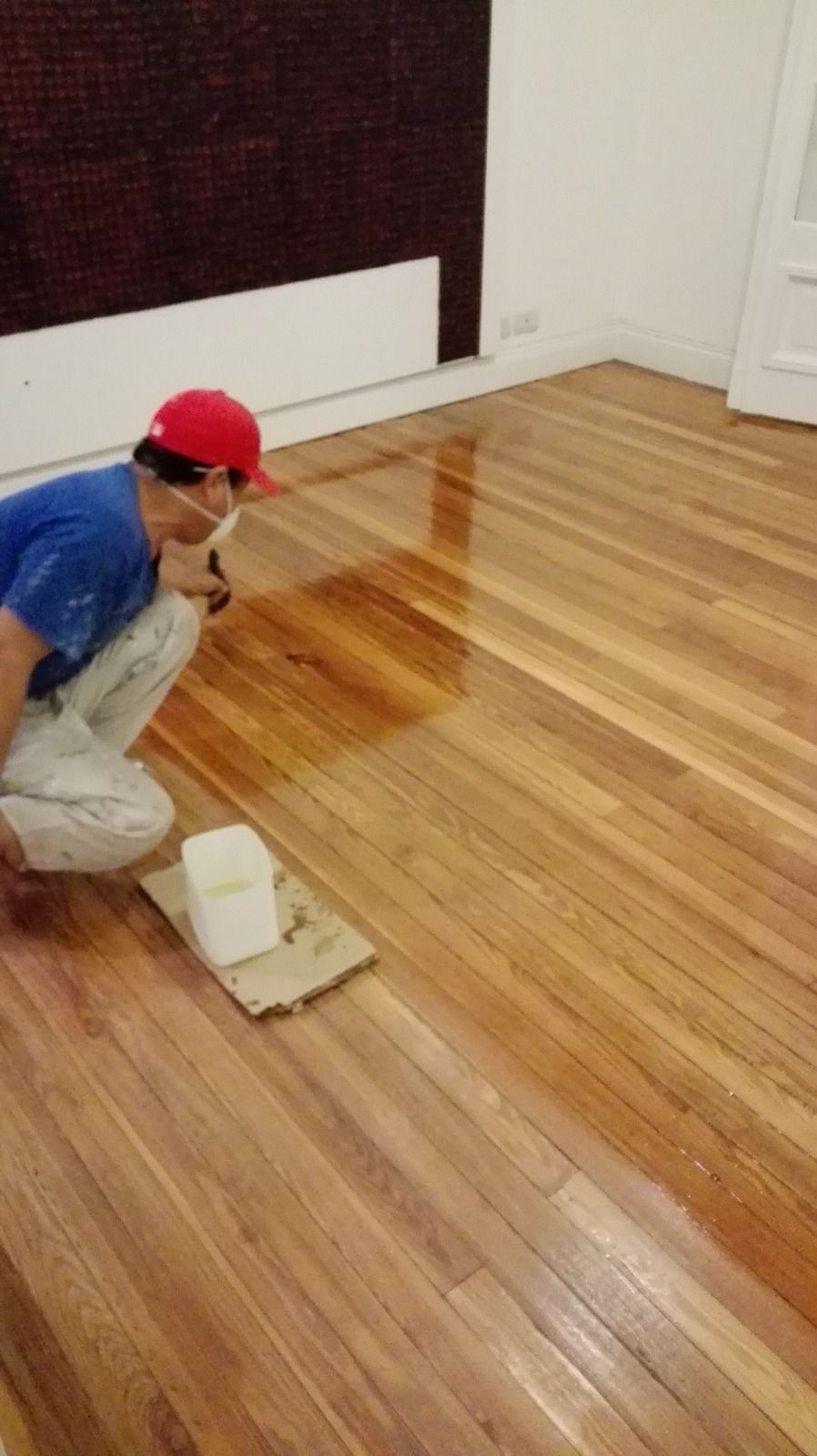 Un trabajador con gorro rojo restaura un suelo de madera en una habitación vacía.