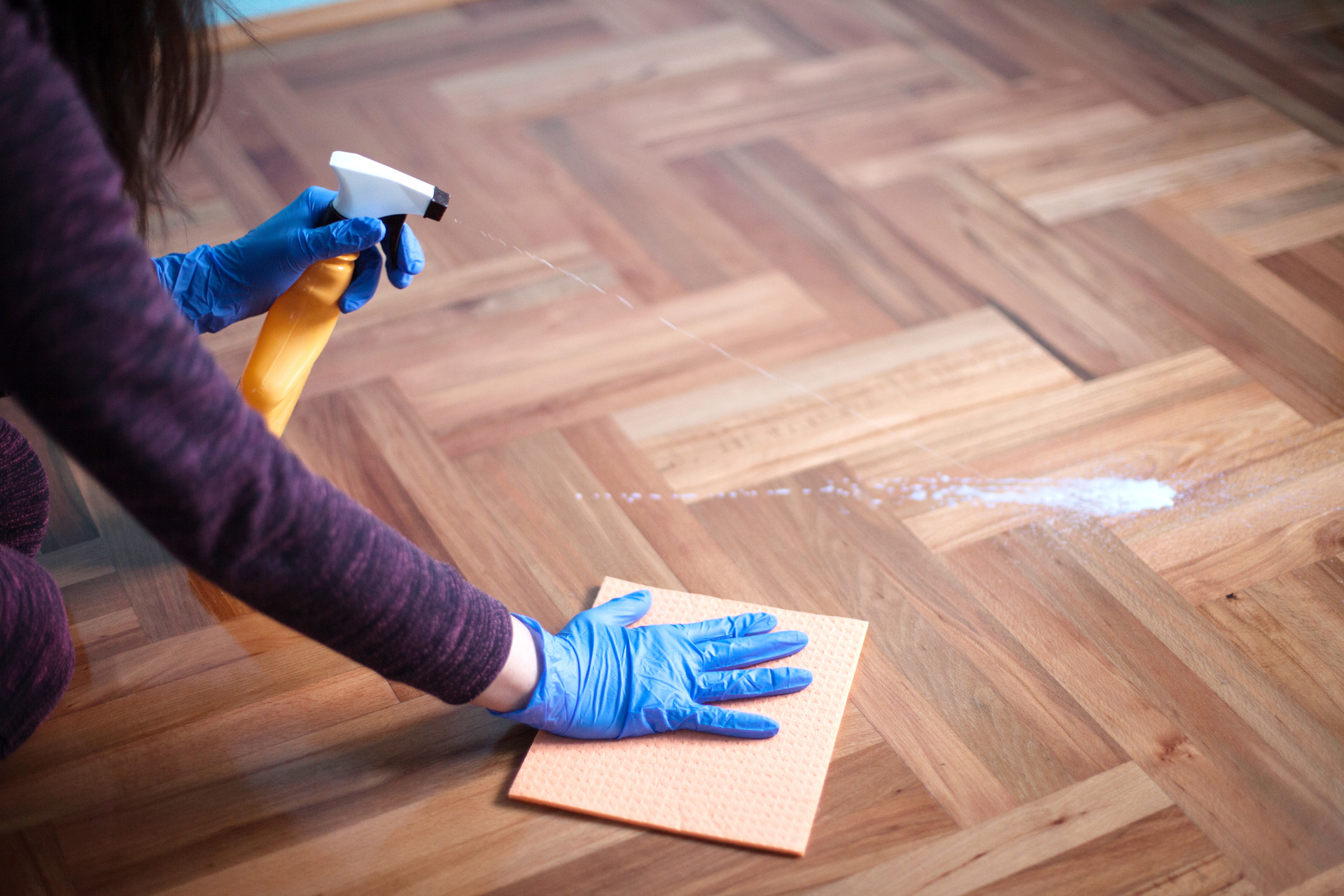 A woman is cleaning a wooden floor with a spray bottle and a cloth.