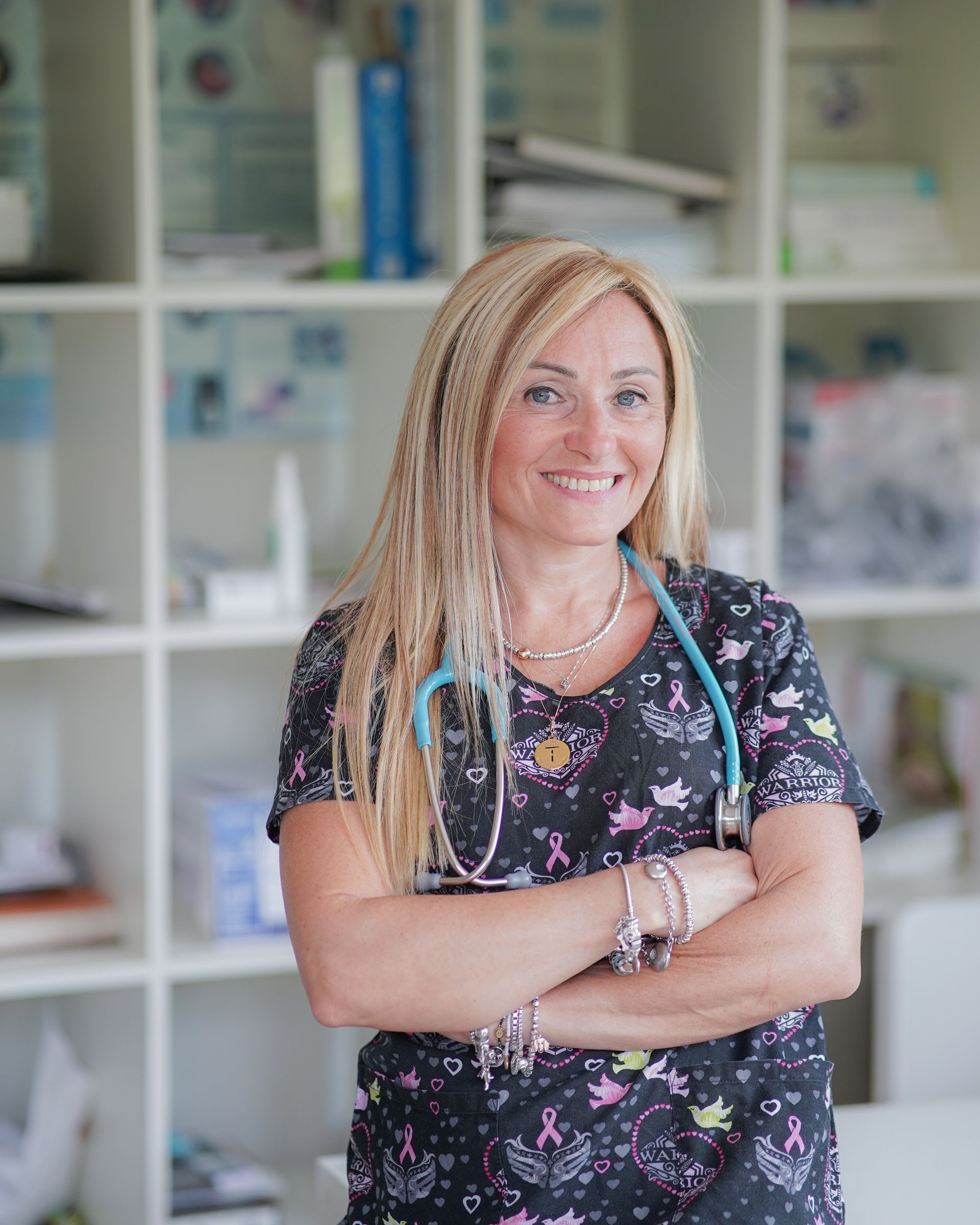 Donna con i capelli biondi e lo stetoscopio, braccia incrociate, sorridente in un ambiente clinico.