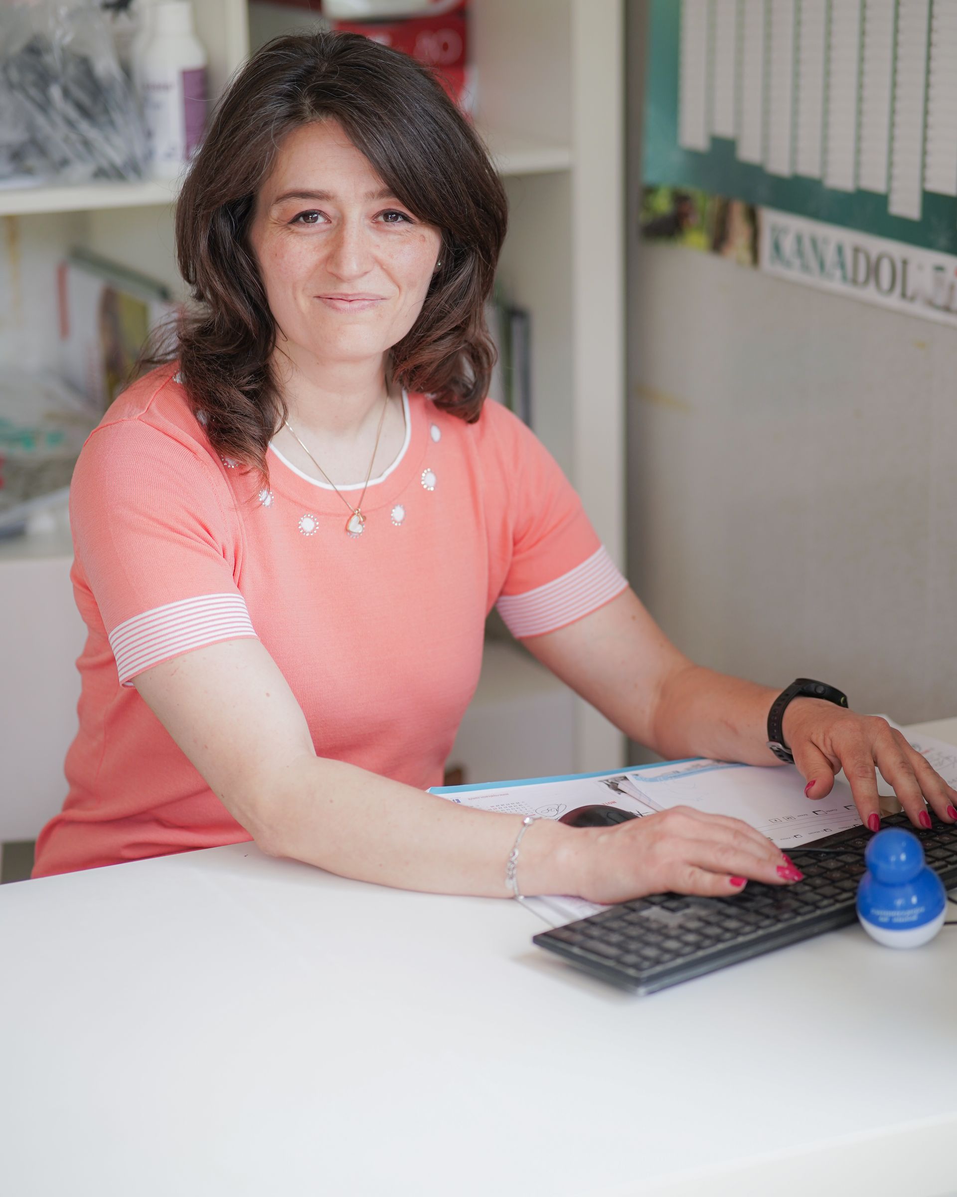 Donna seduta alla scrivania, che scrive. Indossa una camicia color pesca e sorride. Scrivania bianca, libreria sullo sfondo.