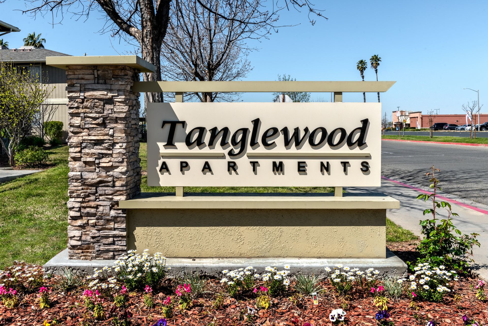 Sign for Tanglewood Apartments, brown and beige stone pillars, tan sign with black lettering, flowers in front.