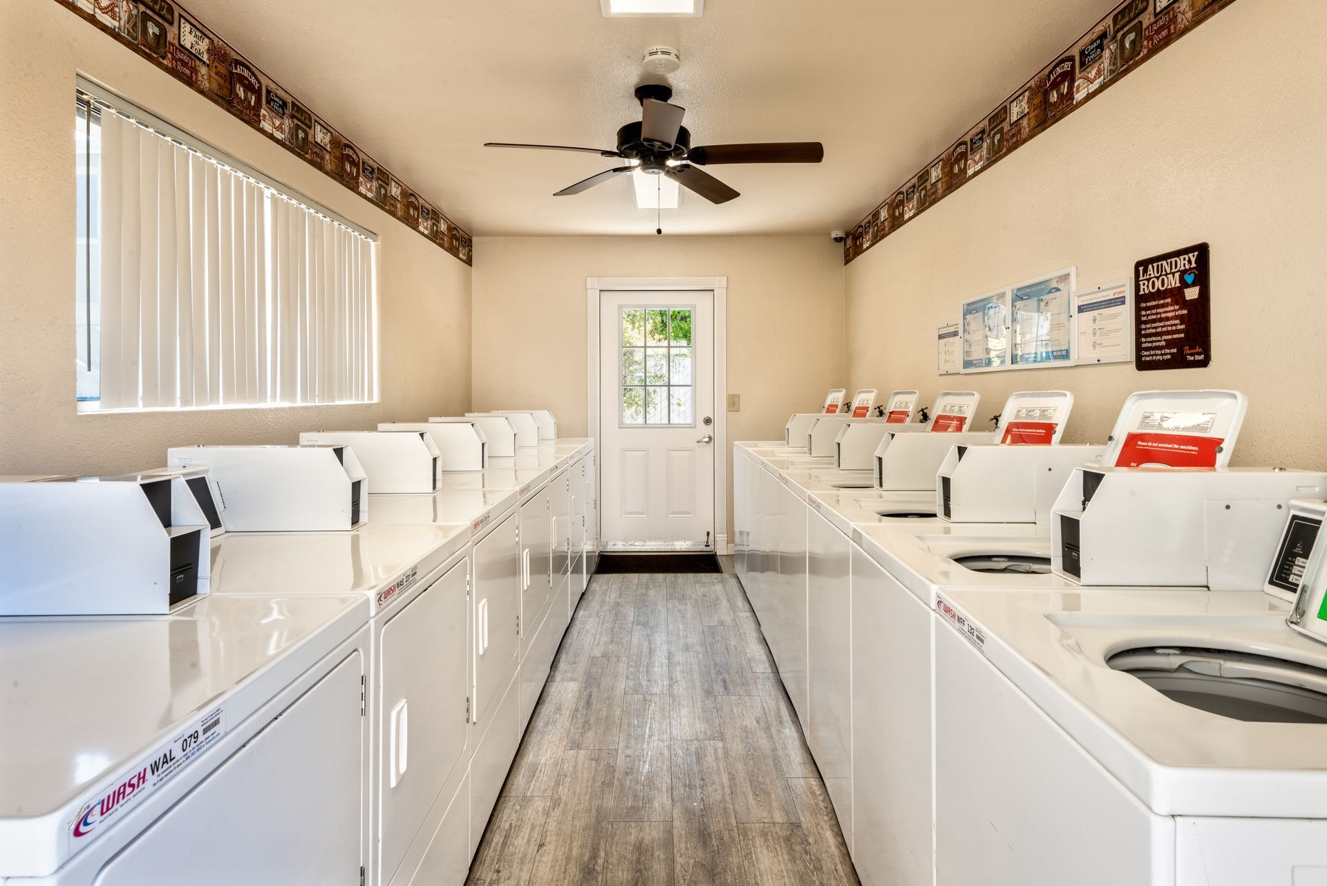 Row of white washing machines in a laundromat, with a door and a window with blinds visible.