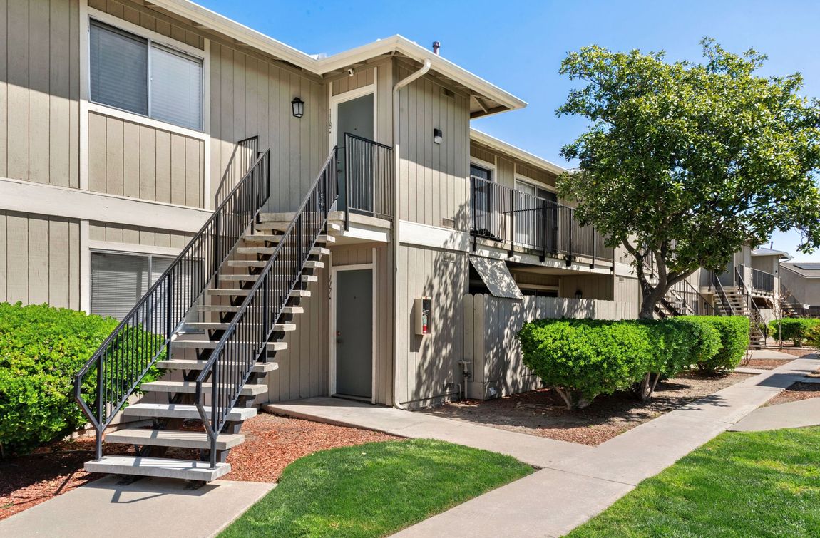 Apartment building exterior, two-story, with stairs, black railings, beige siding, green bushes, and blue sky.